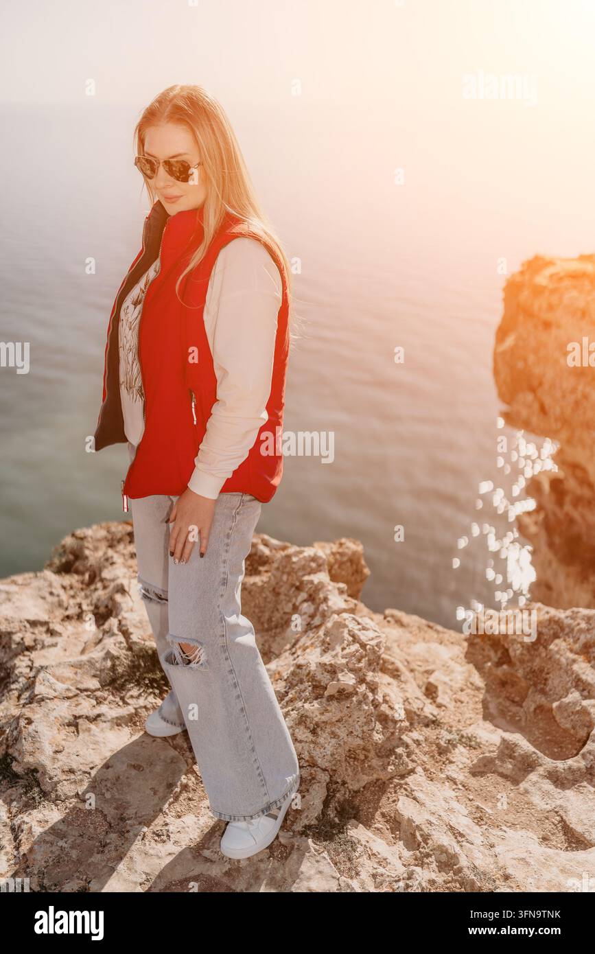 Donna in piedi su un bordo roccioso sulla scogliera che si affaccia sull'oceano, indossa un giubbotto rosso durante una giornata soleggiata di viaggio e avventura Foto Stock