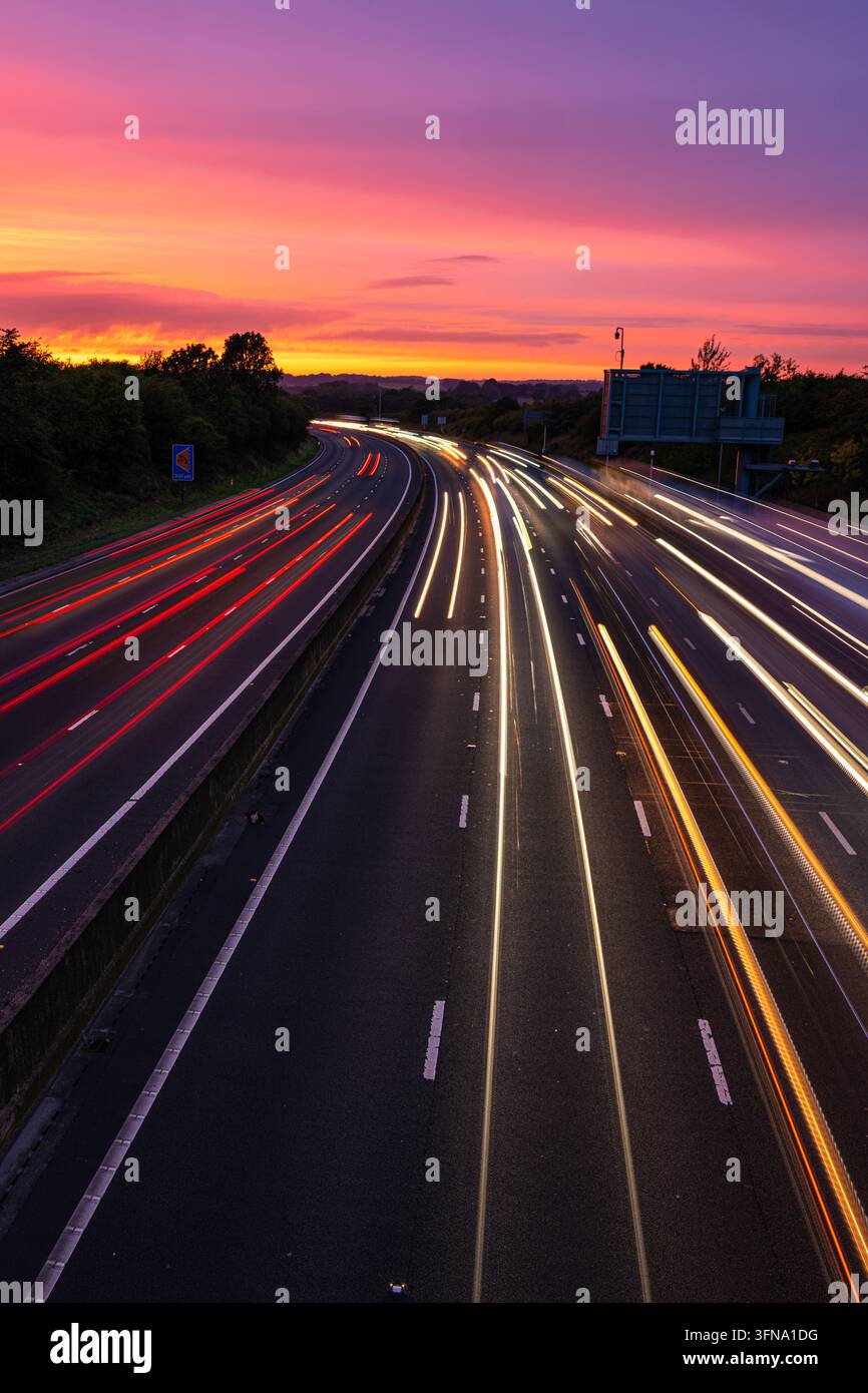 Scie di luci del semaforo a lunga esposizione su un'autostrada trafficata che si curva verso un cielo vivace viola e arancione al tramonto durante l'ora di punta serale. Foto Stock