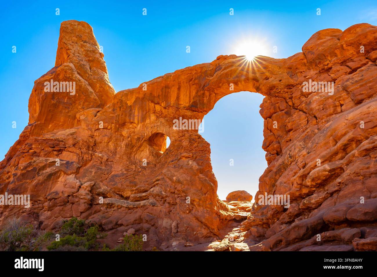Un brillante raggio di sole di mezzogiorno irradia direttamente dietro l'apertura rocciosa rossa e frastagliata di Turret Arch nell'Arches National Park vicino a Moab, Utah Foto Stock