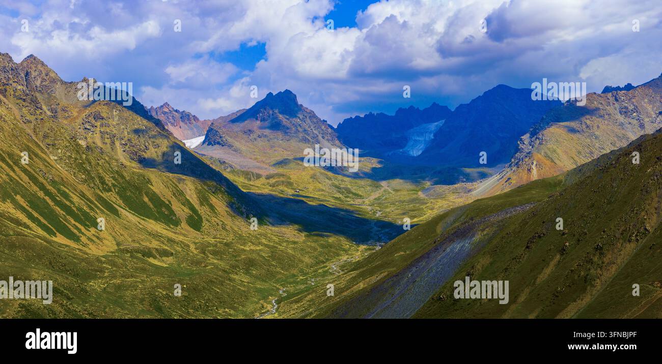 Una vista panoramica cattura una valle montana con un ghiacciaio sullo sfondo. Dolci colline verdi e un ruscello scorrono attraverso il fondo della valle sotto un cielo parzialmente nuvoloso. Foto Stock