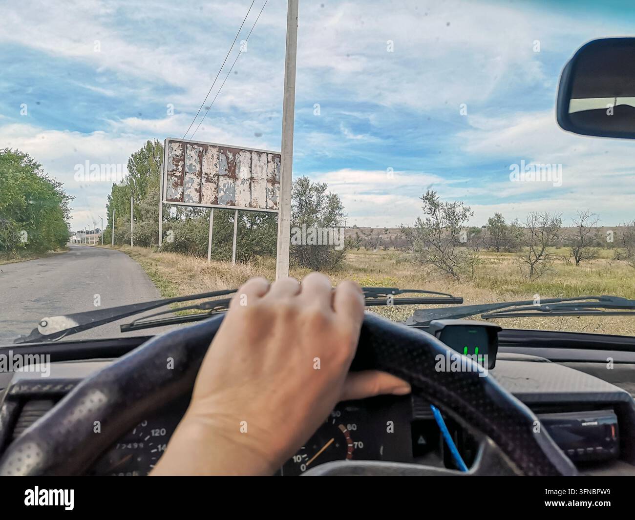 Punto di vista del conducente che tiene il volante su una strada asfaltata oltre un cartellone pubblicitario arrugginito e vuoto nella campagna rurale. Concetto di viaggio in auto e road trip - Immagine stock catturata con smartphone