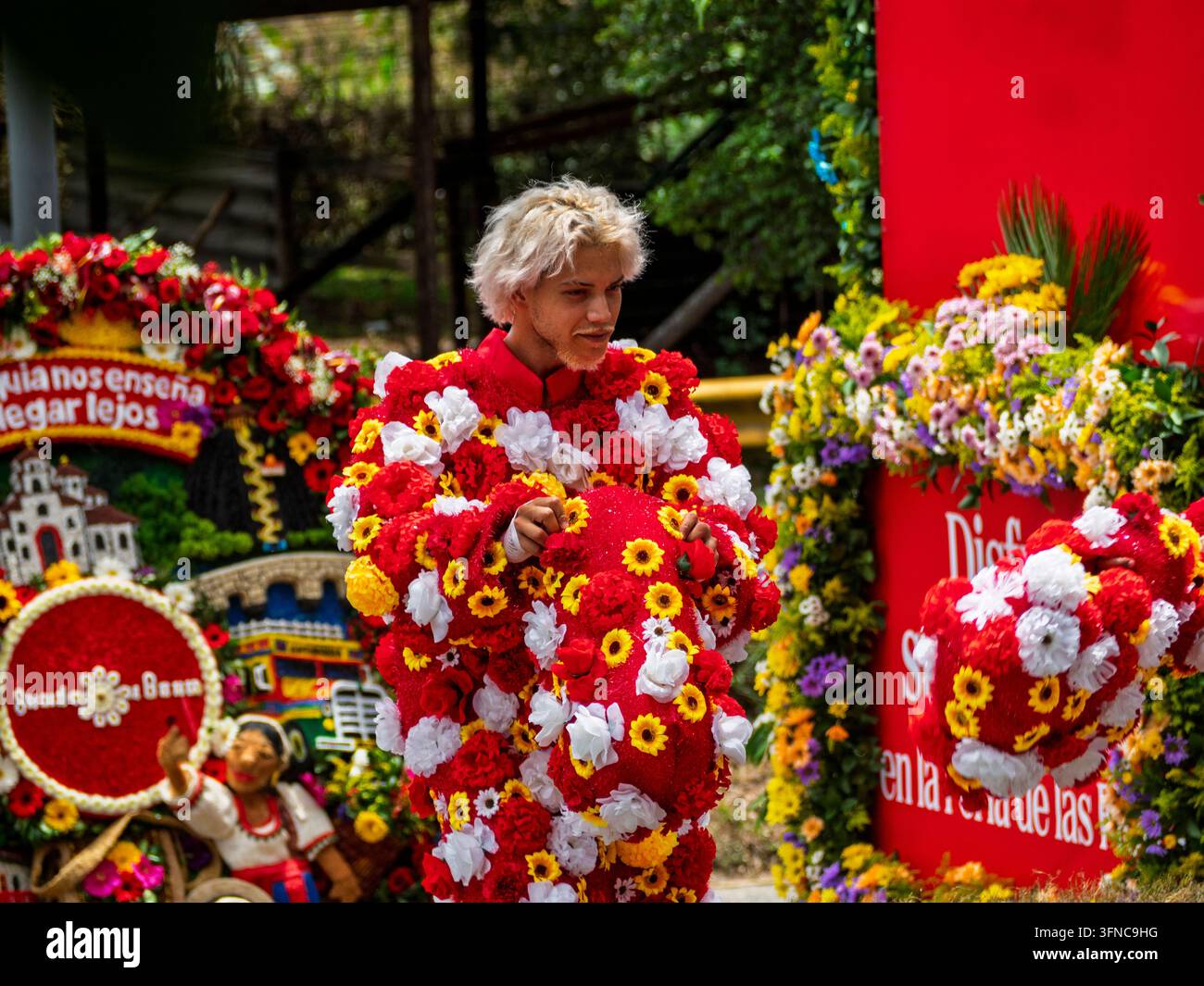 Un performer con capelli biondi decolorati che indossa un costume elaborato ricoperto di fiori finti rossi, gialli e bianchi sorride durante la Feria de las Foto Stock