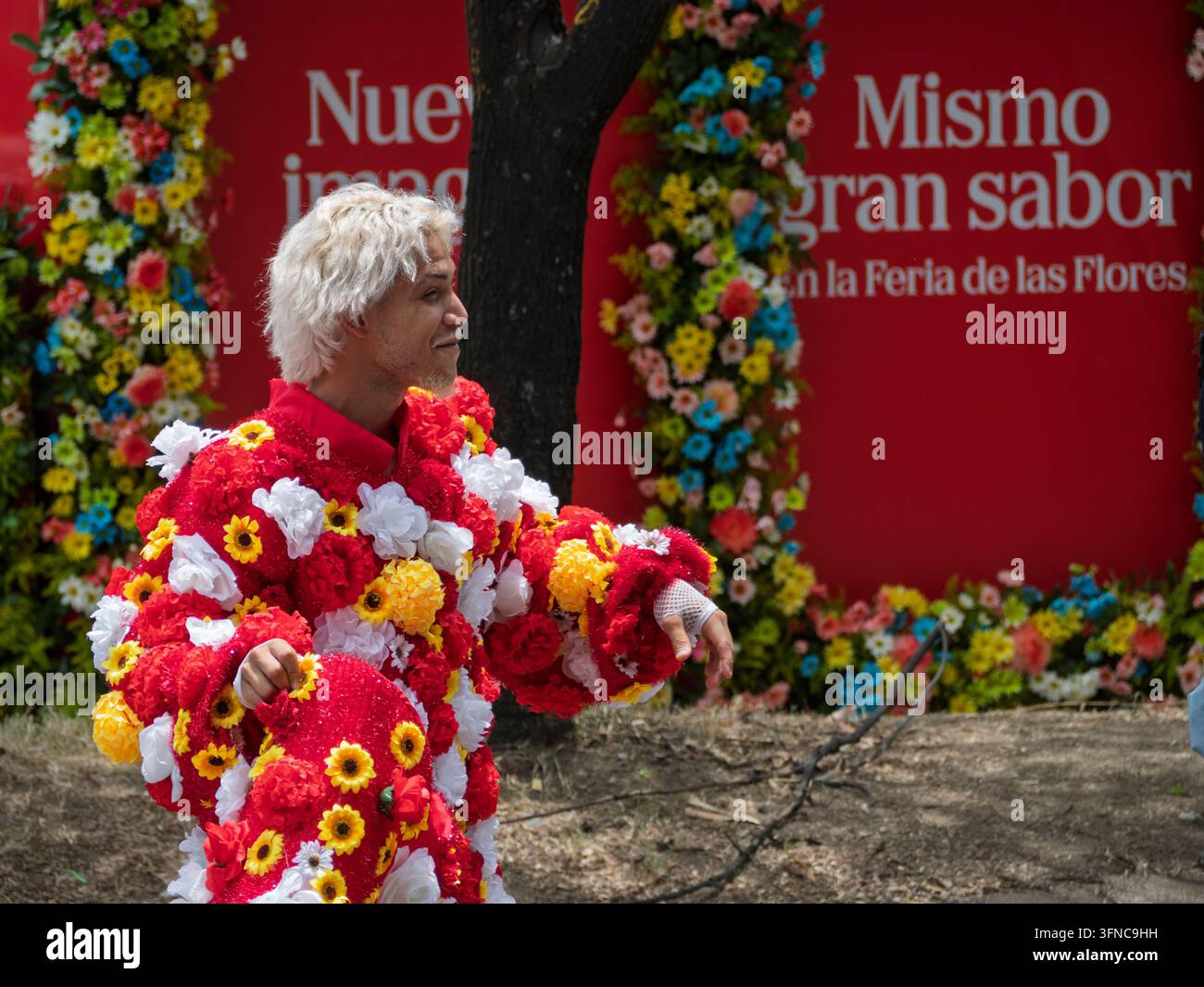 Un performer con capelli biondi decolorati che indossa un costume elaborato ricoperto di fiori finti rossi, gialli e bianchi sorride durante la Feria de las Foto Stock