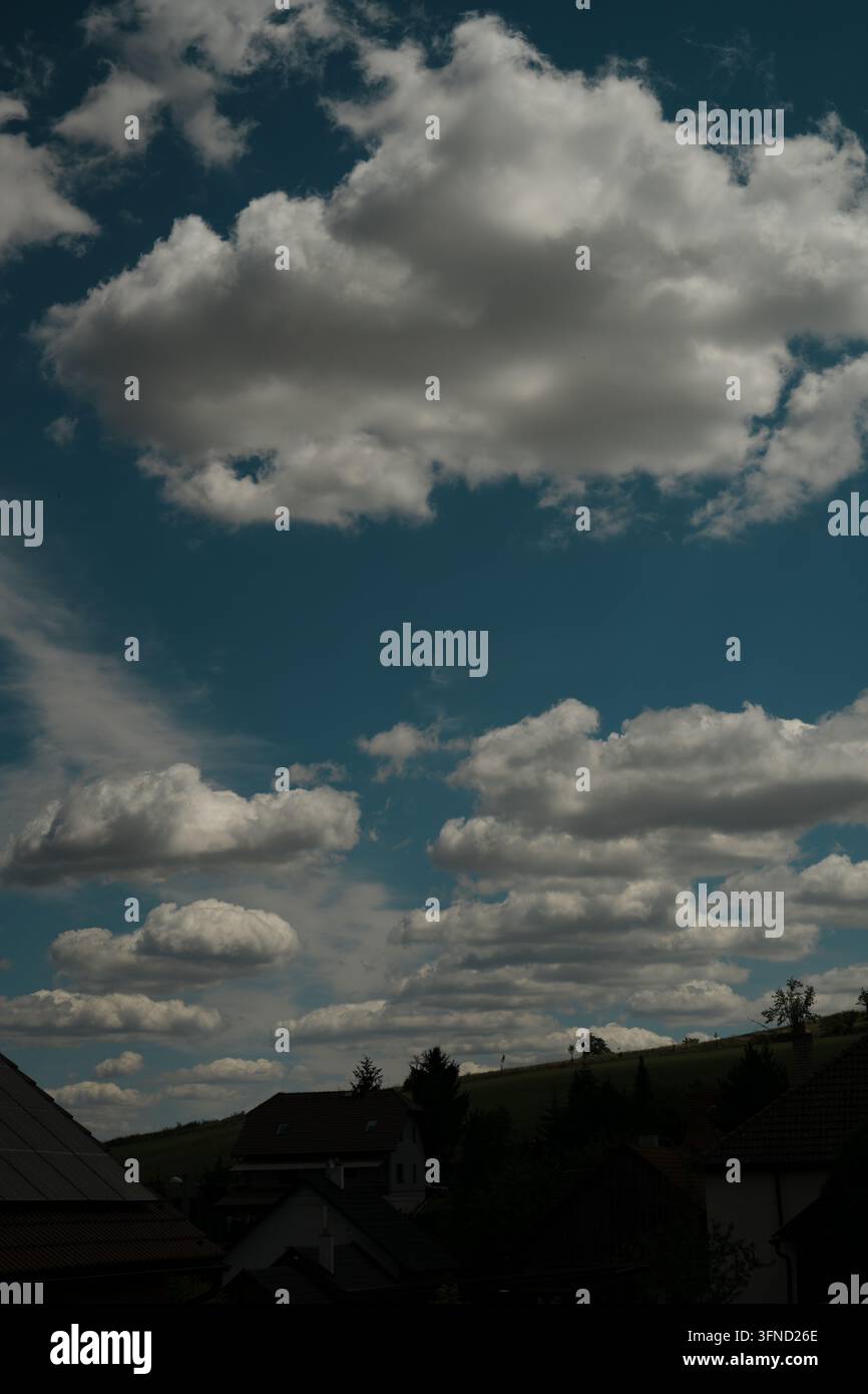 La fotografia mostra un cielo azzurro pieno di nuvole bianche e grigie sopra una linea di alberi e tetti. Edifici con tetti a spiovente sono visibili nella parte inferiore Foto Stock