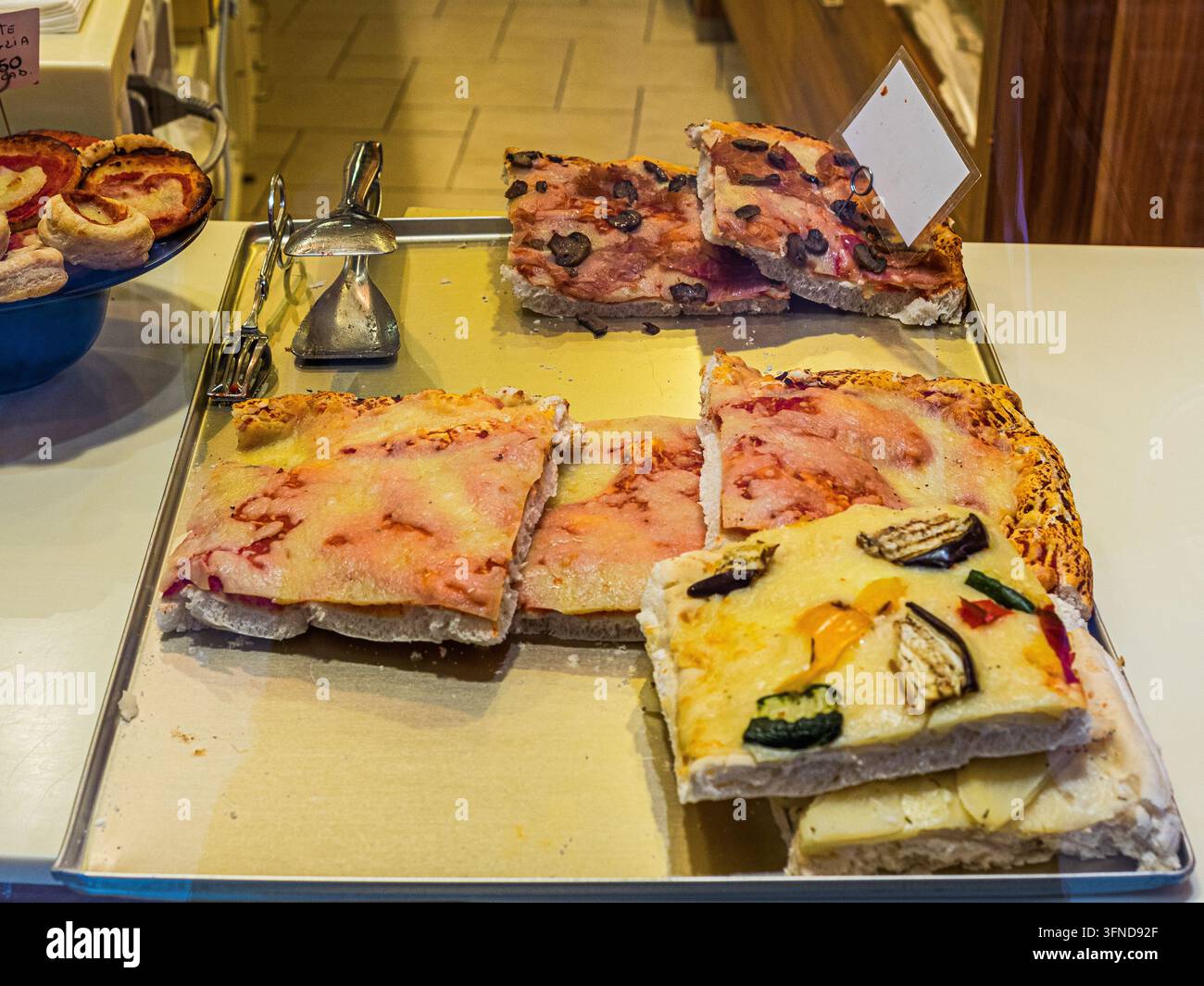 Fette assortite di pizza italiana ricoperte di formaggio, prosciutto, olive e verdure grigliate esposte su un vassoio di metallo in una pizzeria locale. Foto Stock
