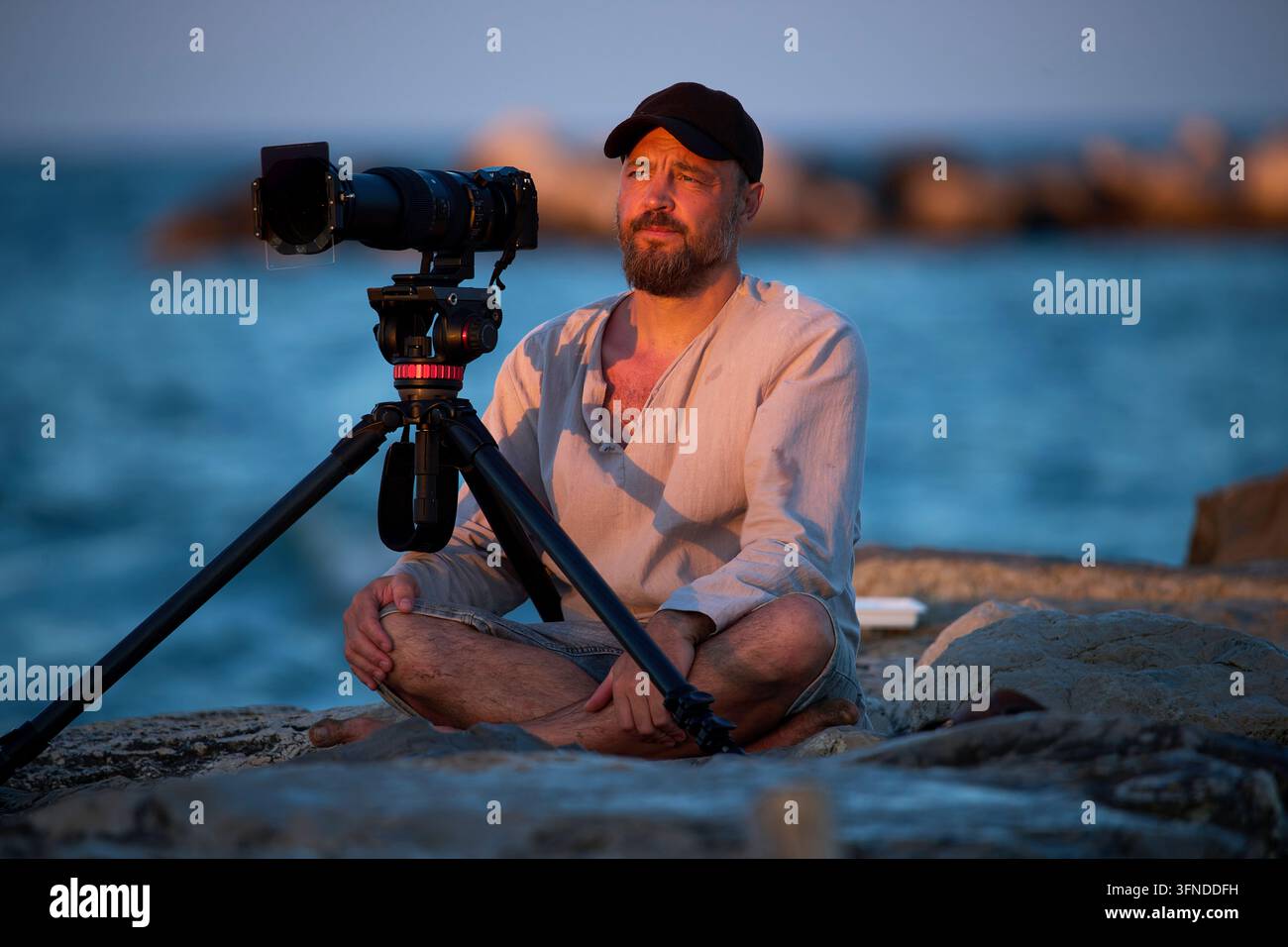 Uomo seduto sul mare con una fotocamera al tramonto Foto Stock