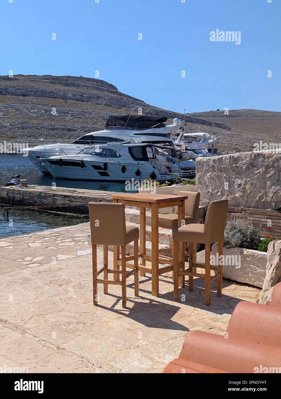 Pirovac, Croazia 2026 Ambientazione all'aperto illuminata dal sole con tavolo alto in legno e quattro sgabelli da bar imbottiti posizionati su un molo di pietra accanto a una marina calma, con acqua azzurra Foto Stock