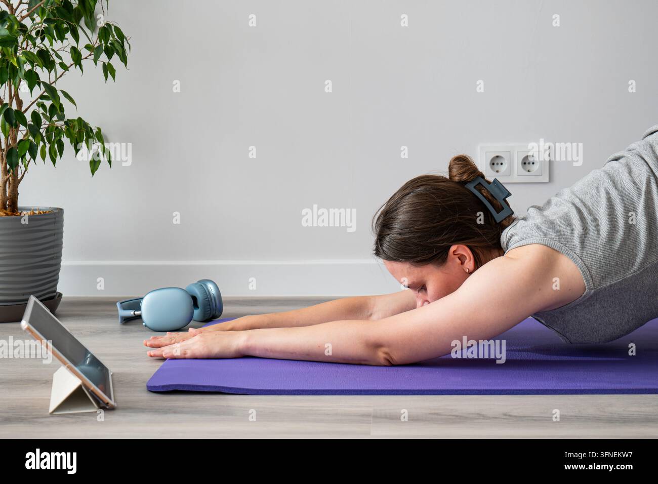 Donna pratica yoga su un tappeto viola. Si piega in avanti con le braccia distese. I suoi capelli sono raccolti, calma e concentrata. Un tablet riposa accanto a lei per guida Foto Stock