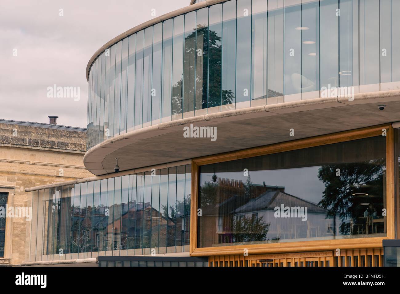Pavimenti in vetro curvi e una finestra con telaio in legno della Blavatnik School of Government a Oxford, che riflette alberi e case, accanto a un edificio in pietra più antico Foto Stock