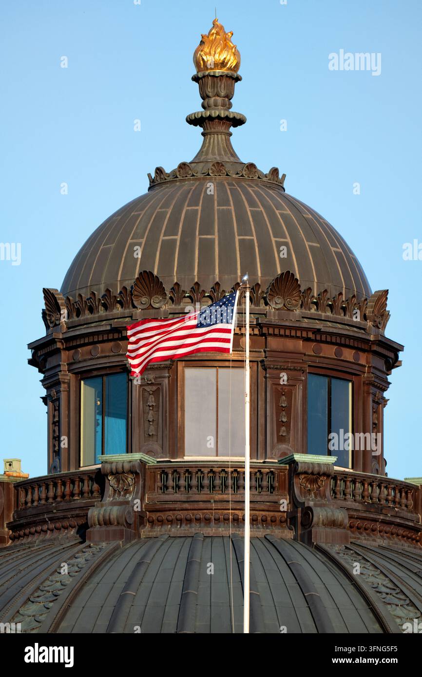 Bandiera in cima alla cupola del Jefferson Building a Washington DC | WASHINGTON — Una bandiera americana sventola al vento in cima al lanternino della cupola del Thomas Jefferson Building della Library of Congress a Capitol Hill. Il candelabro dorato di bronzo della Conoscenza sopra di esso sormonta la struttura a circa 195 piedi (59 m), un faro simbolico scelto per segnalare lo scopo dell'edificio come deposito di conoscenza piuttosto che di potere, in contrasto deliberato con la cupola del Campidoglio nelle vicinanze. Foto Stock