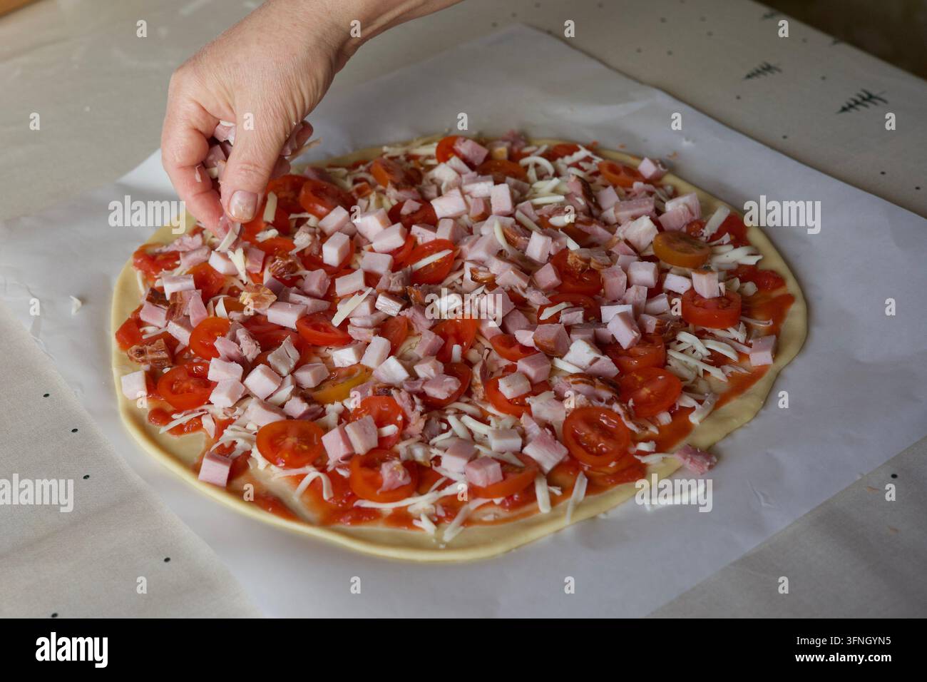 Mano che aggiunge guarnizioni a cubetti a una pizza fatta in casa cruda con formaggio e fette di pomodoro. Foto Stock
