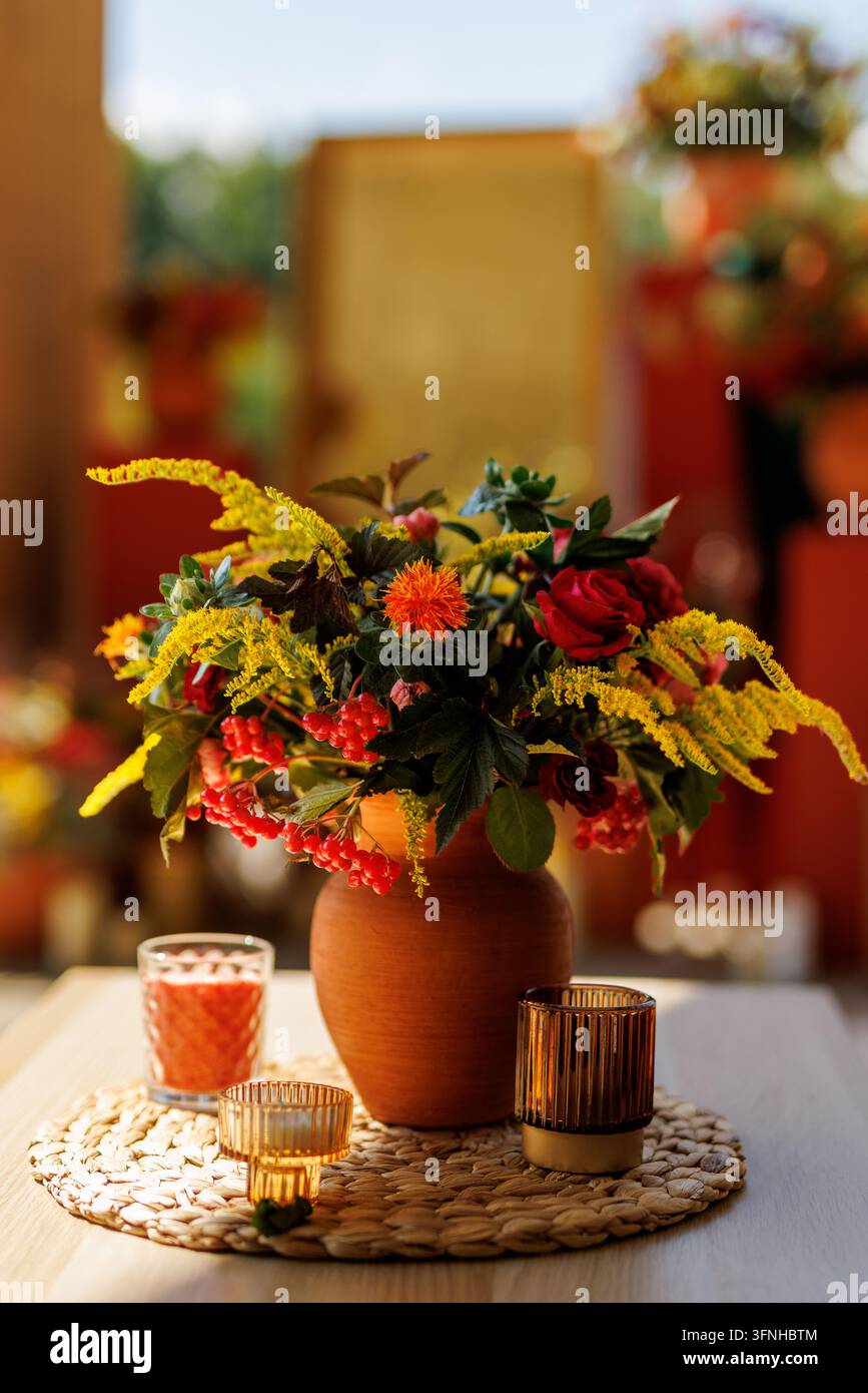Decorazione da tavolo centrale con rose rosse, fiori gialli e bacche in un vaso di terracotta, affiancato da portacandele in vetro. Foto Stock