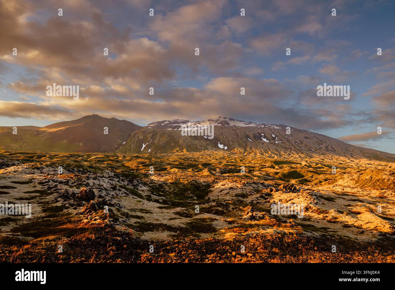 La luce dorata del tramonto cade su un paesaggio montano accidentato con stratovulcano ricoperto di ghiacciai, neve a chiazze, terreno roccioso e nuvole drammatiche sotto un Foto Stock