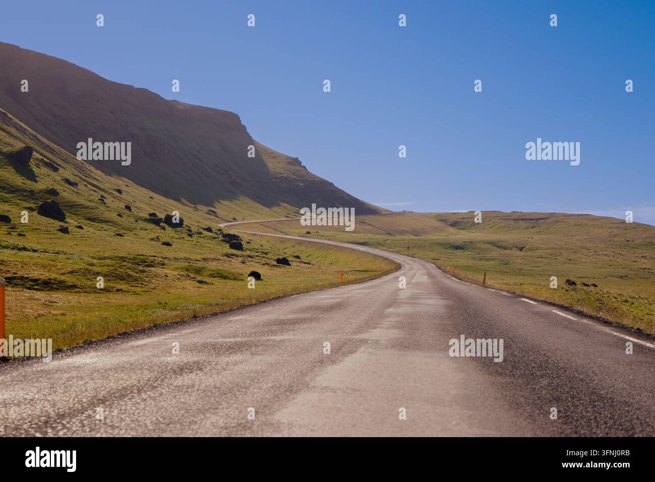 Strada asfaltata tortuosa e vuota Snæfellsnesvegur attraverso colline montuose verdi sotto un cielo blu limpido, con pendici rocciose e una campagna aperta e vasta. Foto Stock