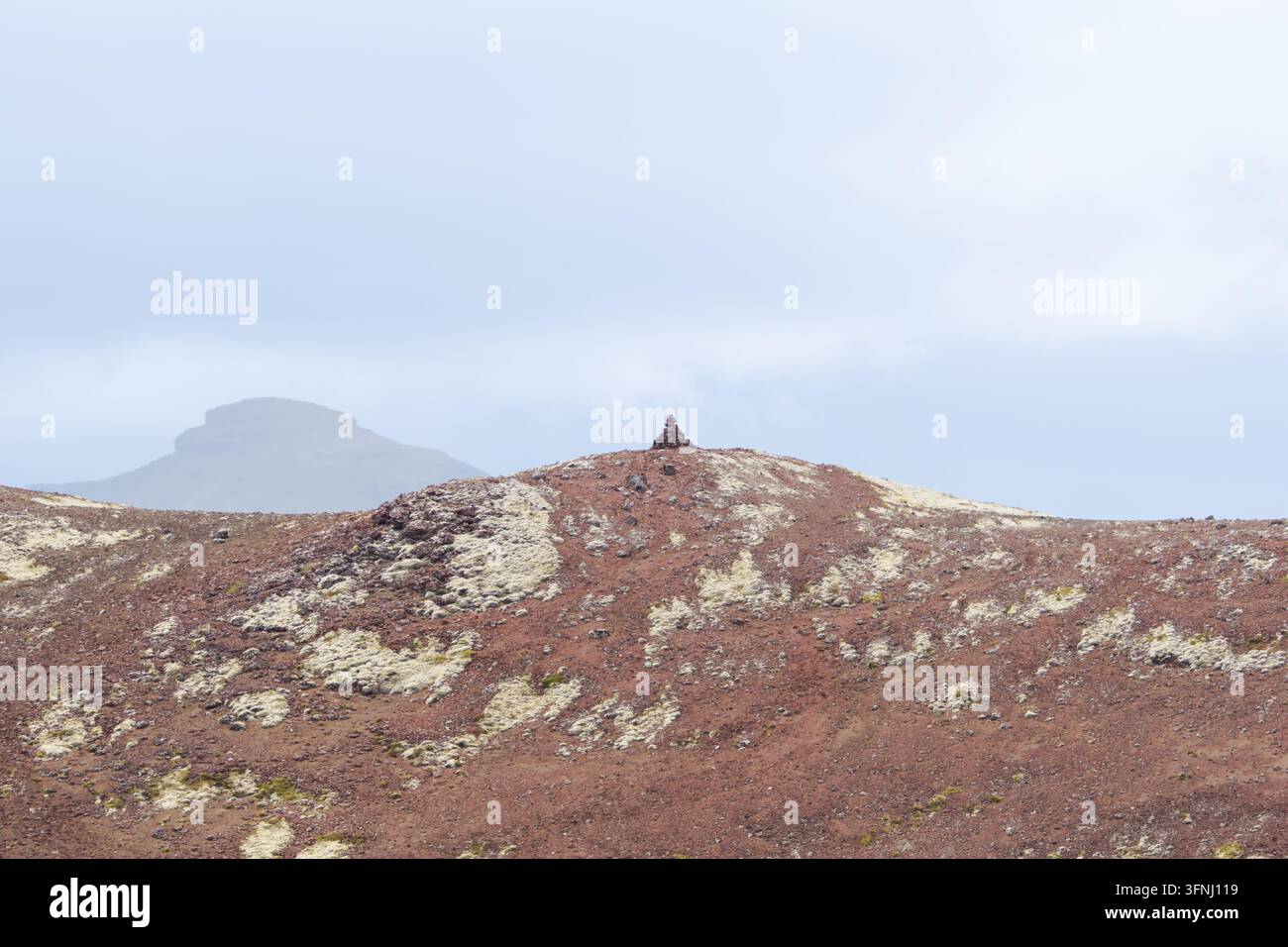 Un piccolo cumulo di pietre si erge su un pendio roccioso rosso coperto di muschio pallido, con una montagna nebbiosa che svanisce in un cielo grigio e soffice nei fiordi occidentali dell'Islanda Foto Stock