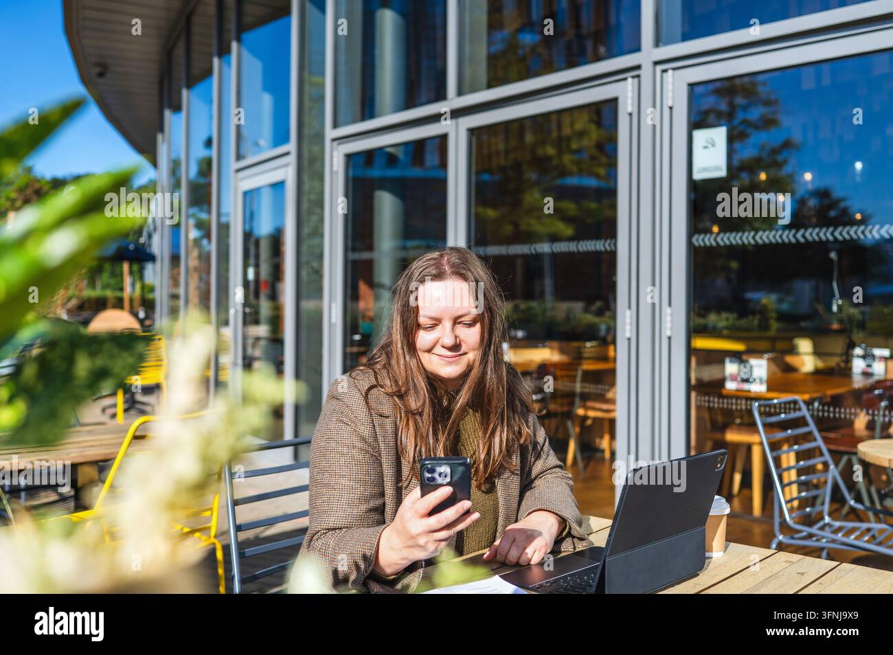 Una freelance utilizza uno smartphone e un laptop mentre è seduta a un tavolo all'aperto di un caffè, rappresentando il lavoro remoto, la comunicazione digitale e la produzione aziendale Foto Stock