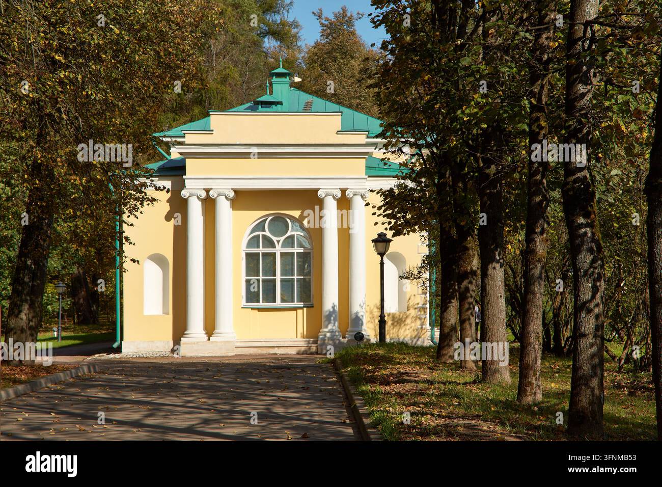 Padiglione giallo con colonne bianche e una finestra ad arco si trova accanto a un marciapiede lastricato, circondato da alberi maturi e foglie d'autunno cadute Foto Stock