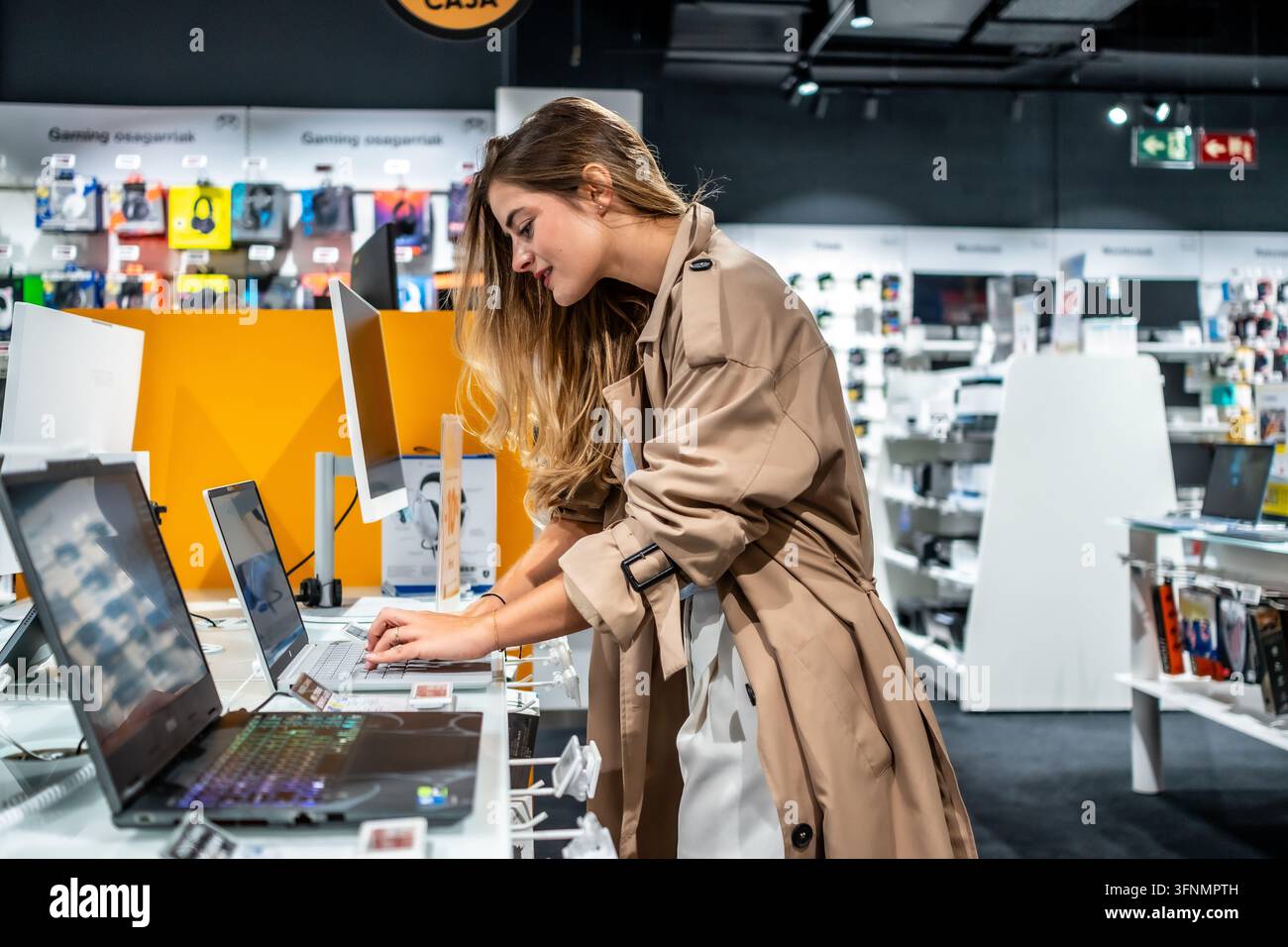 Giovane donna in un trench coat in piedi in un negozio di elettronica moderno, che testa i laptop in esposizione e valuta le caratteristiche per prendere una decisione di acquisto mentre Foto Stock