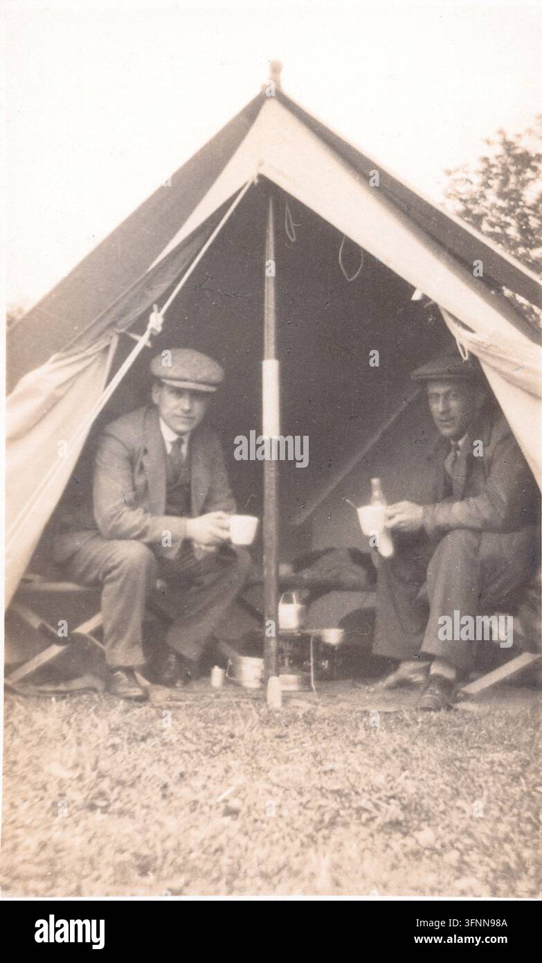 Scena di campeggio dei primi del XX secolo che mostra due uomini seduti all'interno di una tenda a cresta. La fotografia cattura un campo ricreativo all'aperto con arredi semplici e attrezzature da cucina, che riflette il campeggio ricreativo e le attività di vacanza prima della Prima Guerra Mondiale. Da una foto pubblicata per la prima volta da Thomas Illingworth & Co. di Londra intorno al 1910. Foto Stock