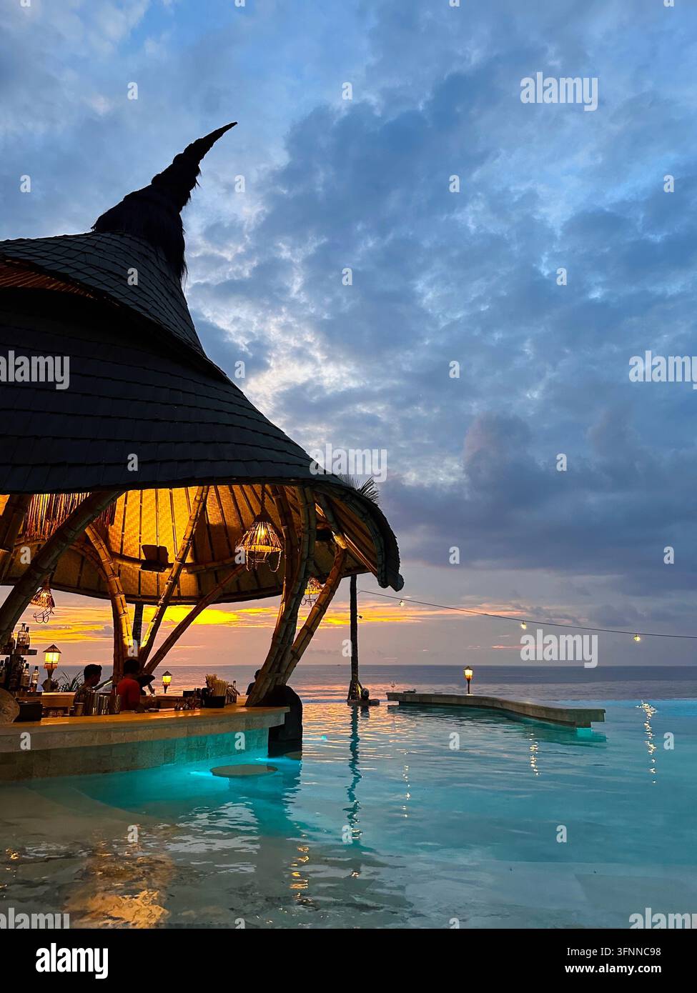 Bar della piscina in bambù illuminata si affaccia sull'oceano sotto un drammatico cielo al tramonto. - Immagine stock catturata con smartphone