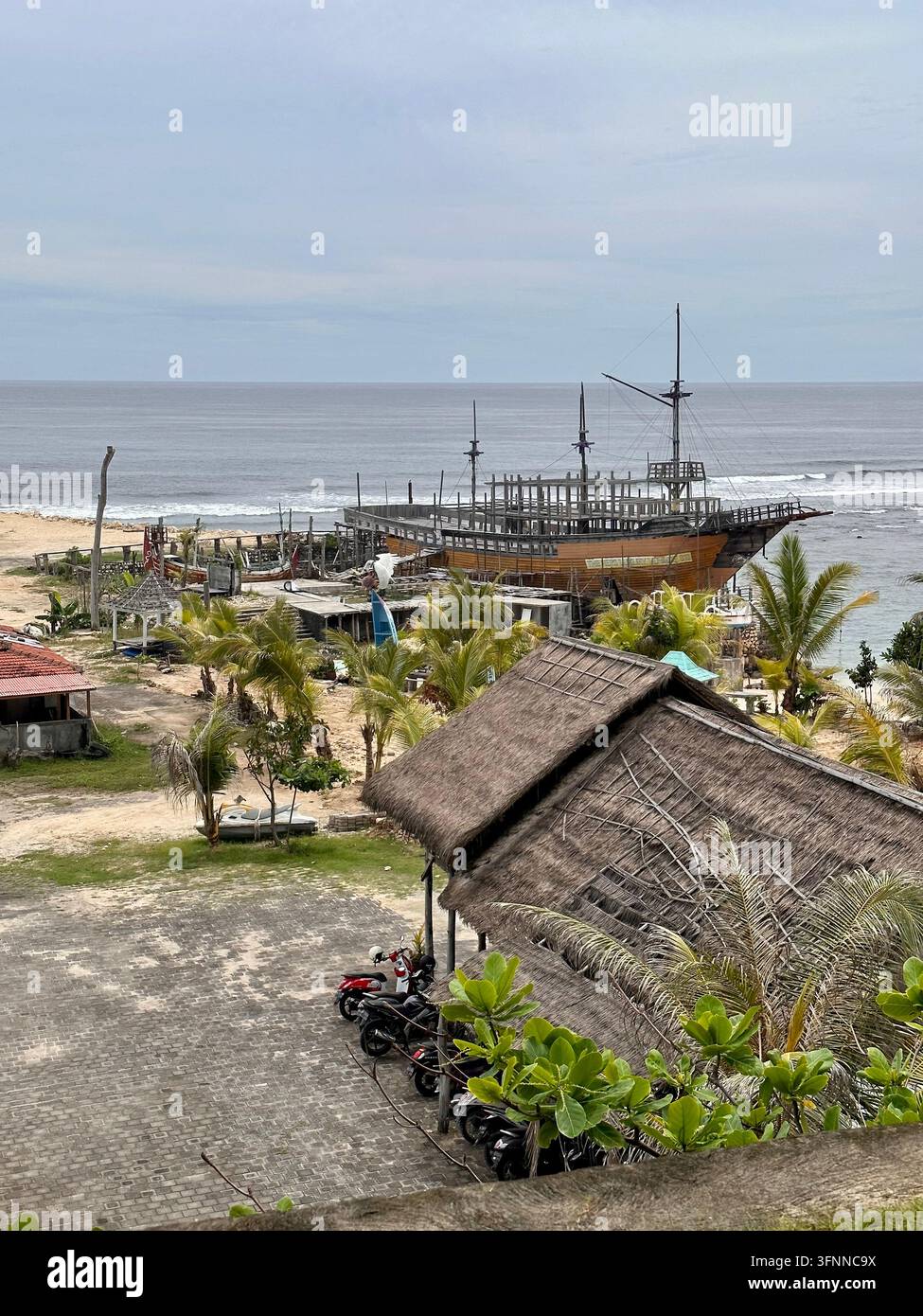 Struttura in legno incompiuta di una nave si affaccia sull'oceano accanto ad edifici con tetto di paglia in una luce diurna nuvolosa. - Immagine stock catturata con smartphone