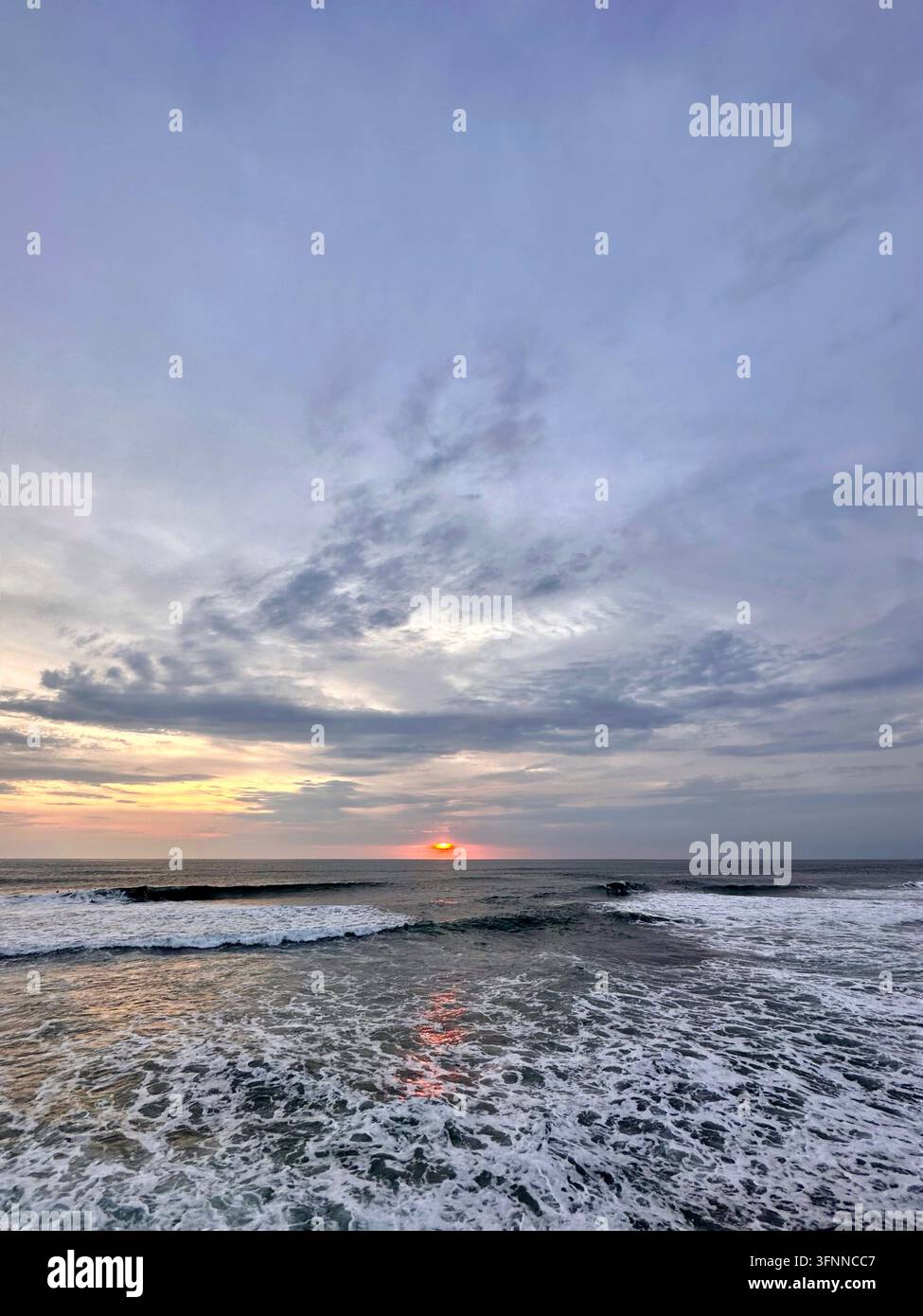 Il sole che tramonta risplende sopra le onde schiumose dell'oceano sotto nuvole serali stratificate. - Immagine stock catturata con smartphone