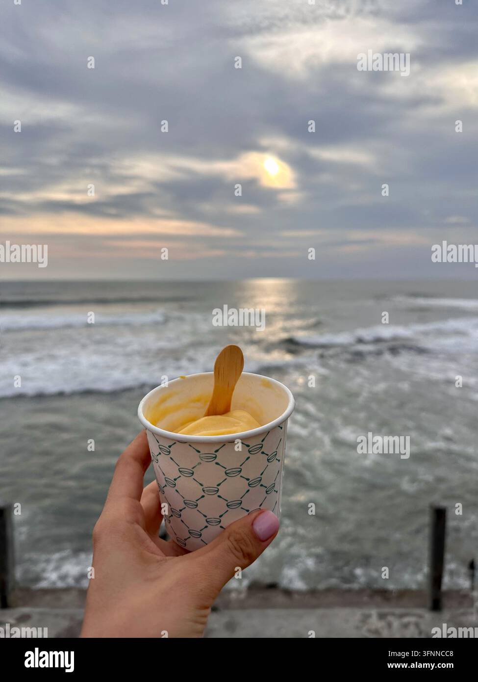Una mano tiene una coppetta di gelato accanto alle onde dell'oceano sotto la luce soffusa del tramonto. - Immagine stock catturata con smartphone