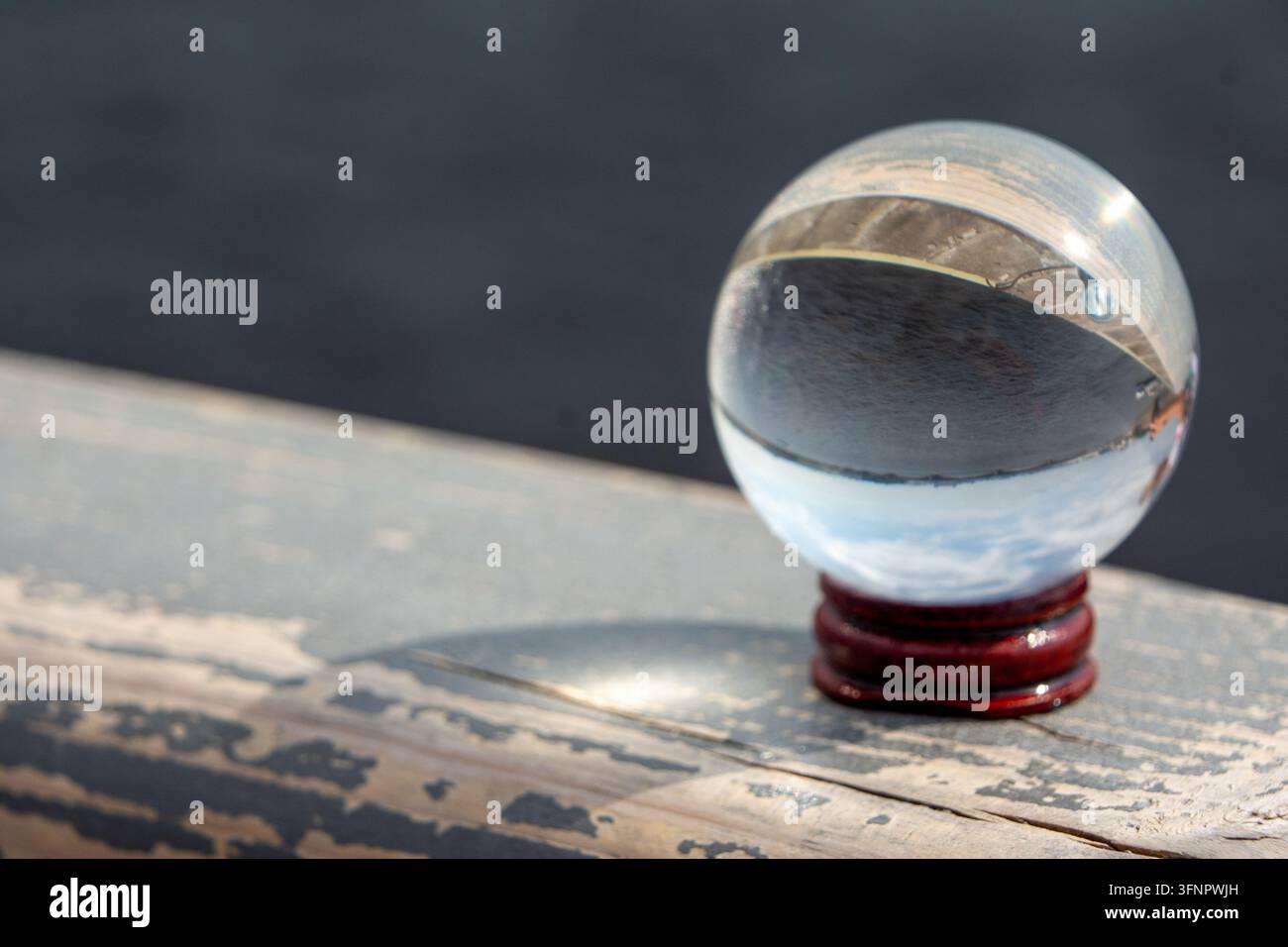 Foto creativa in primo piano di una sfera di vetro trasparente posizionata su un piccolo supporto in legno su una ringhiera o cornicione in legno lungo un fiume. La sfera di cristallo Foto Stock