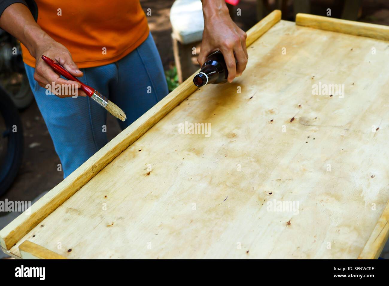 Una persona applica un trattamento per il legno su una tavola di compensato con pennello e bottiglia all'aperto, mostrando una preparazione pratica di falegnameria e costruzione manuale Foto Stock