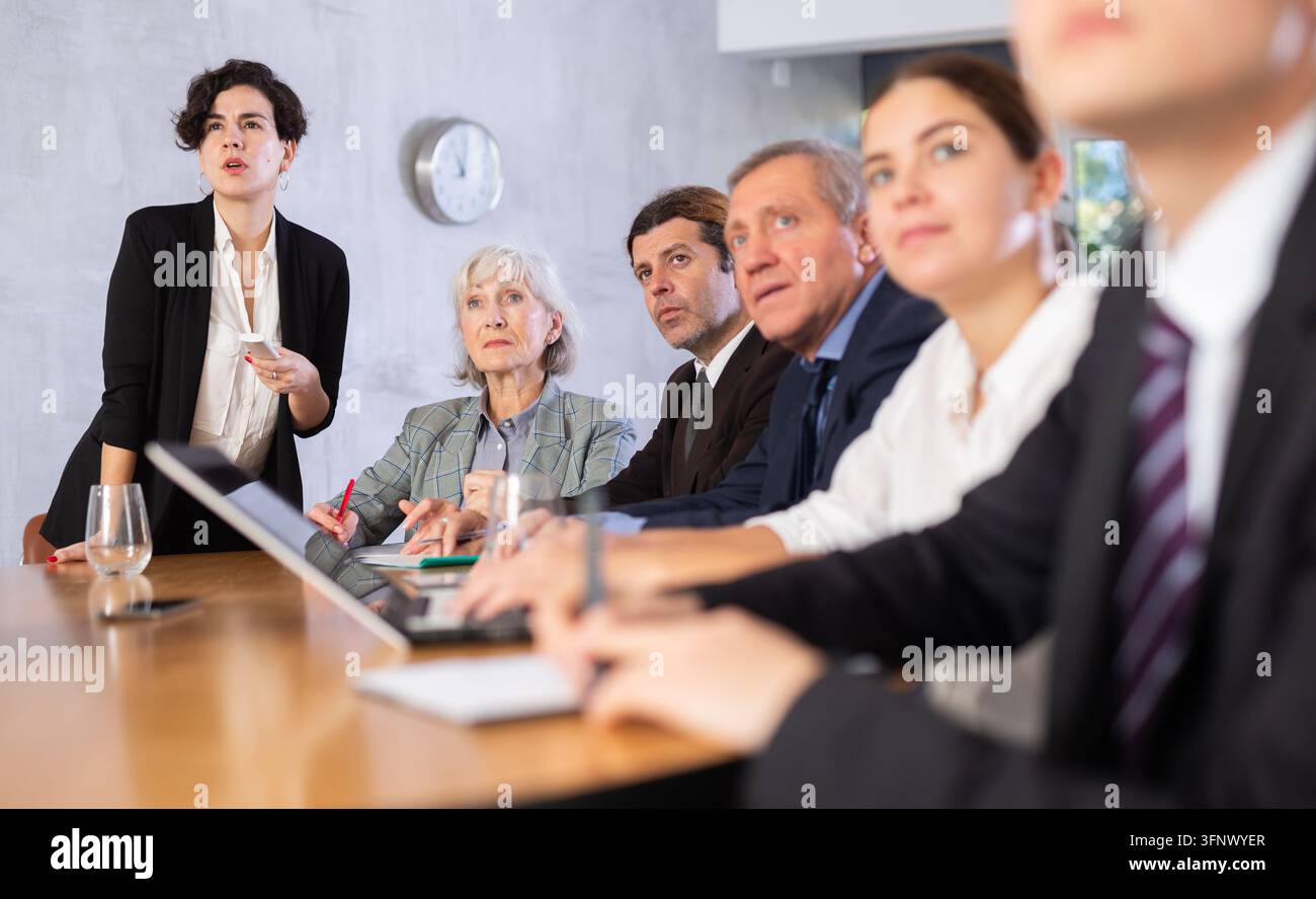 Giovane donna d'affari ispanica sicura di sé e interessata che tiene una presentazione ai colleghi durante una riunione di lavoro in ufficio, utilizzando un telecomando per passare alle diapositive Foto Stock