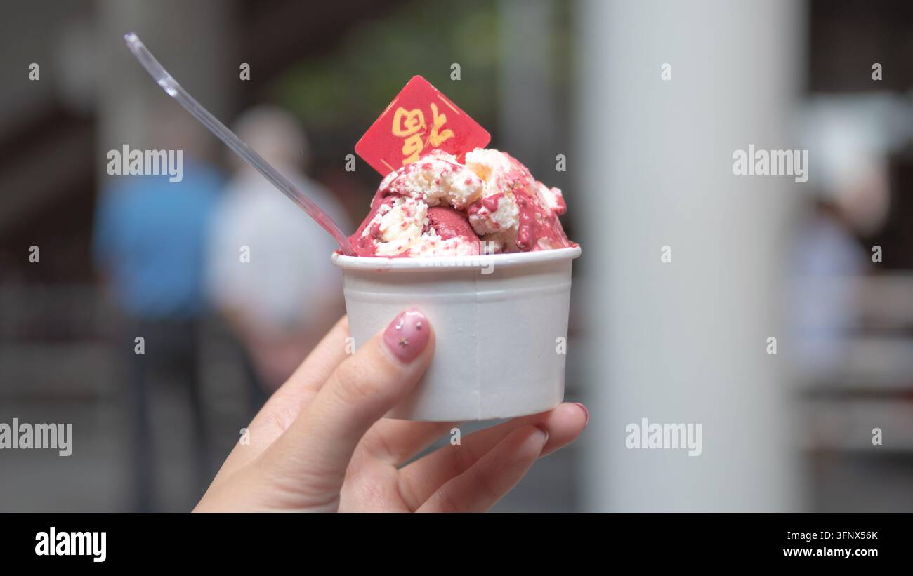 Primo piano di una mano che tiene una tazza di dessert congelato rosa guarnito con panna e una decorazione festiva rossa a Singapore Foto Stock
