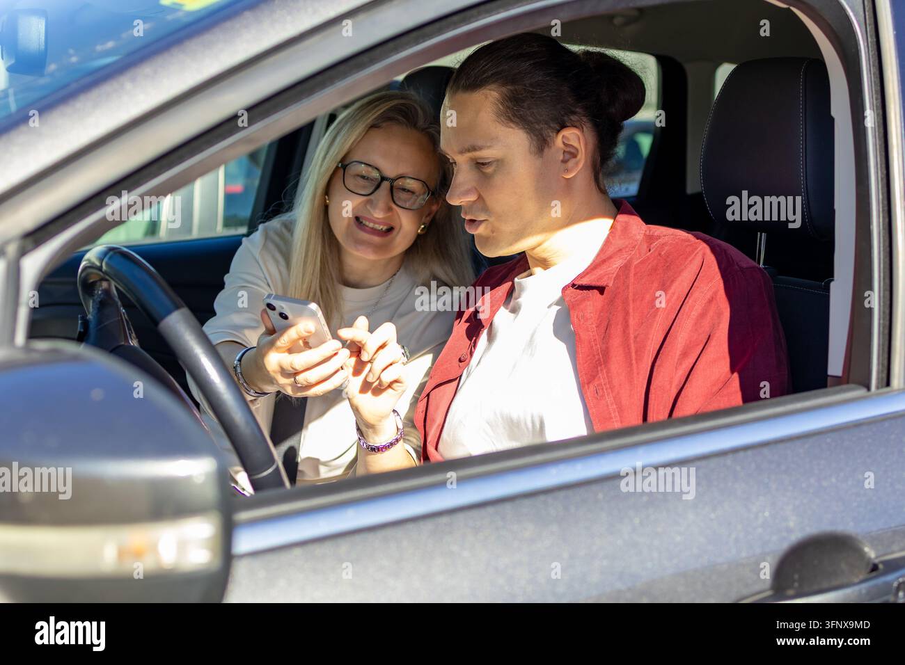 Donna bionda sorridente con occhiali si china verso un uomo in camicia rossa seduto al volante mentre guardano insieme uno smartphone. Concetto di piano Foto Stock