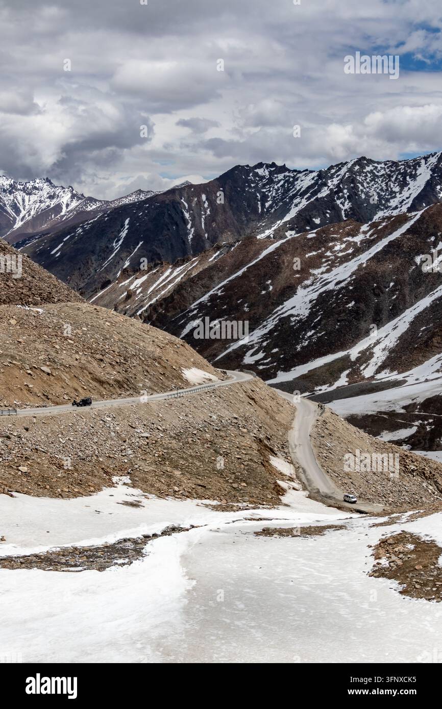 Strada di montagna tortuosa che attraversa una valle di altopiano arida con neve a chiazze, pendii rocciosi ripidi, picchi lontani e un cielo nuvoloso drammatico. Foto Stock