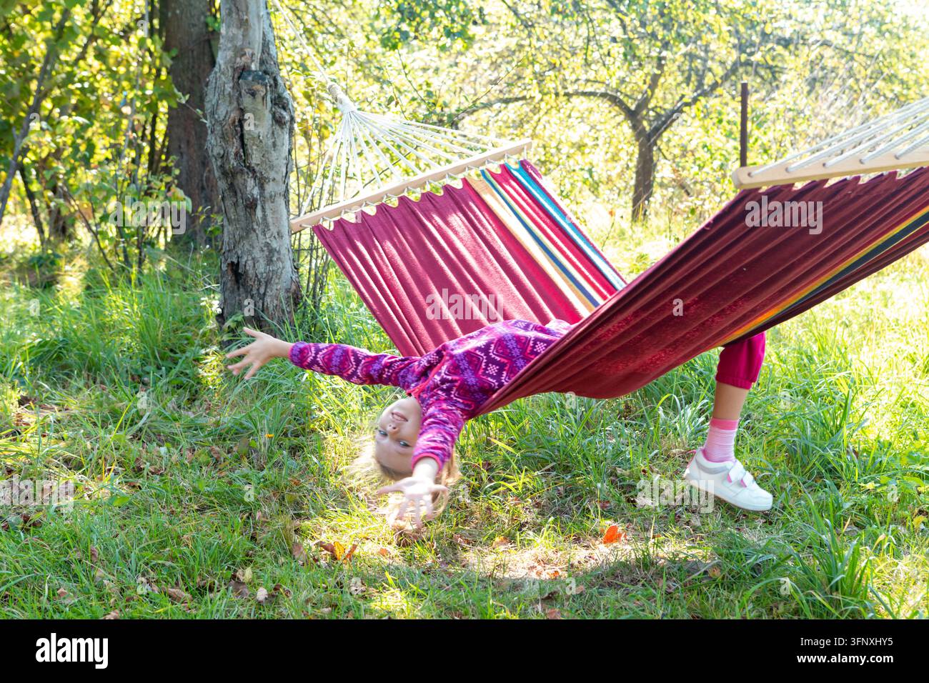 Bambino gioca mentre si rilassa in un'amaca sotto un cielo soleggiato in un parco tranquillo durante il giorno Foto Stock