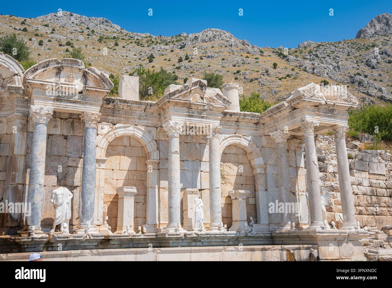 Una vasta veduta frontale dell'antica città di Sagalassos in Turchia mostra superfici di pietra consumate e forme scultoree danneggiate. Un terreno roccioso si innalza dietro la struttura Foto Stock
