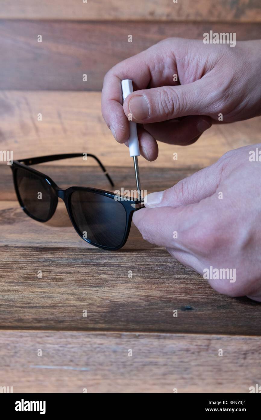 Mani di una persona che regolano una vite su occhiali da sole neri con un mini cacciavite su un tavolo di legno Foto Stock