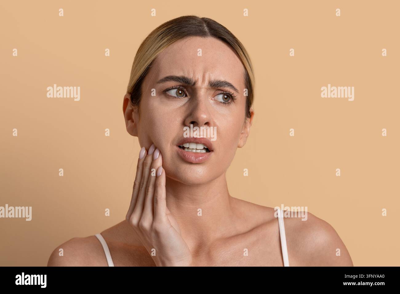 Giovane donna che tocca la mascella con espressione preoccupata su uno sfondo studio beige Foto Stock