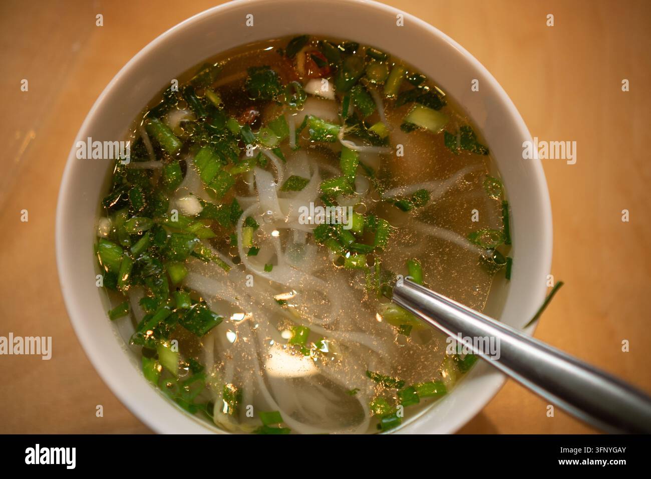 Ciotola bianca di zuppa di brodo chiaro con noodles e cipollotti si trova su tavolo da pranzo in legno, offrendo un'esperienza di cibo confortante e caldo. Un rituale alimentare sano porta pace e soddisfazione in qualsiasi ambiente domestico accogliente Foto Stock