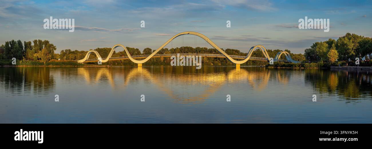 Vista panoramica del ponte pedonale futuristico Wave nel parco Natalka, nel distretto di Obolon a Kiev al tramonto, con la struttura a forma d'onda riflessa nella calma dell'acqua Foto Stock