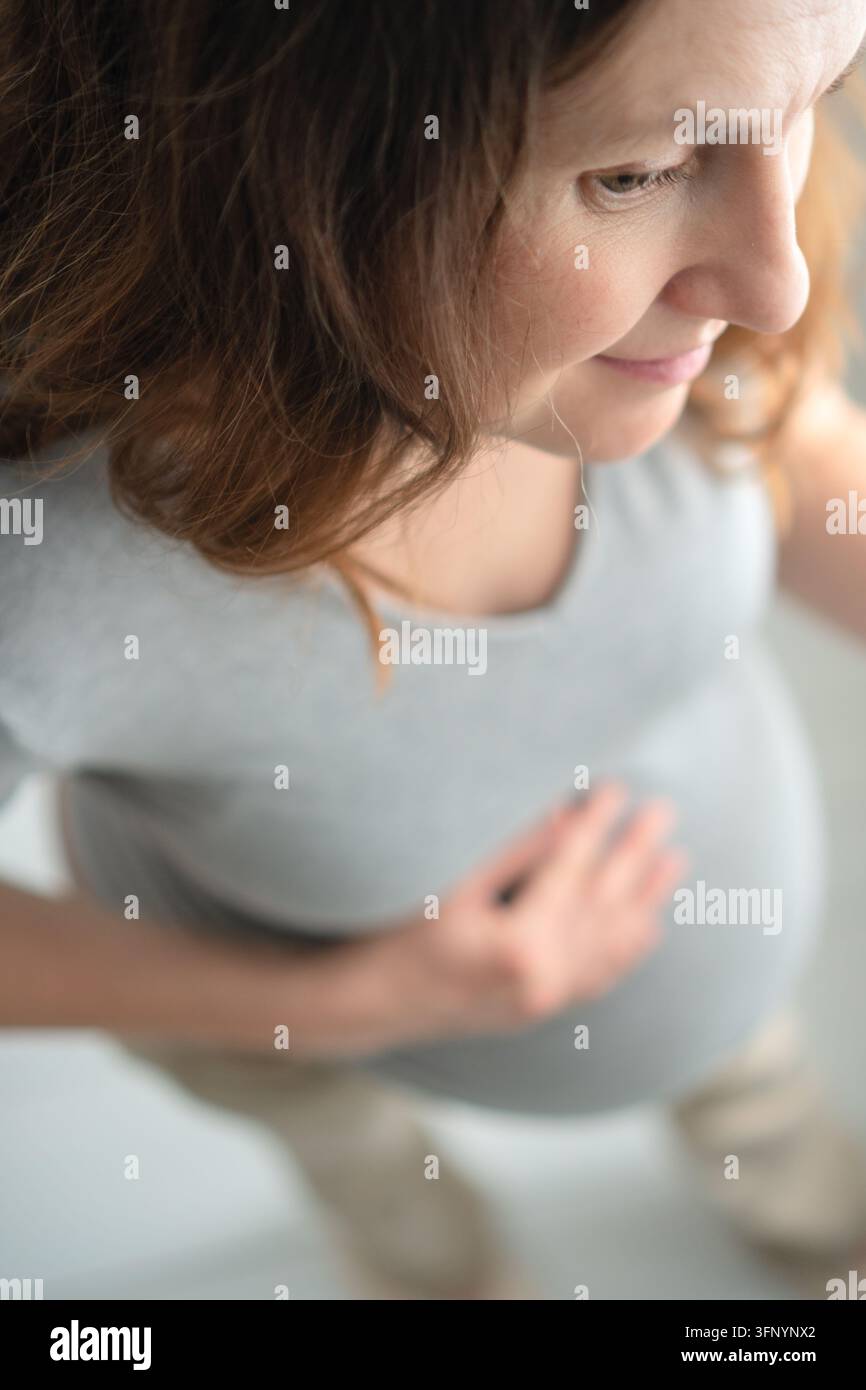 Una donna incinta di profilo riflette mentre tiene il ventre nella luce soffusa. La madre in attesa che guarda verso il basso presenta un momento pensieroso e riflessivo di riflessione materna e contemplazione calma. Foto Stock