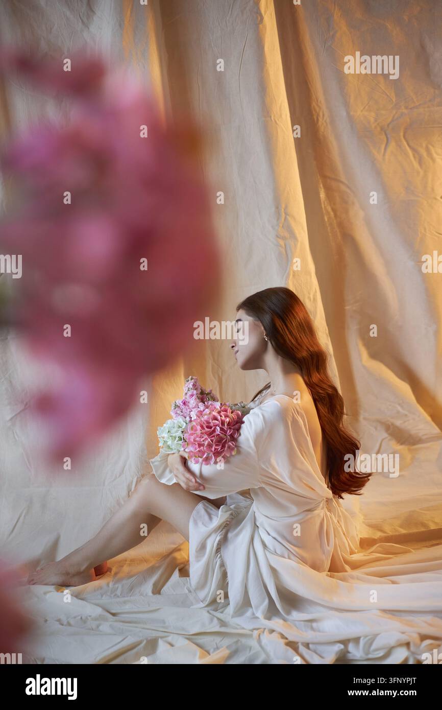 la donna è seduta sul pavimento, tiene un mazzo di fiori. Ha i capelli lunghi e indossa un abito fluido. Una luce soffusa illumina la scena, crea Foto Stock