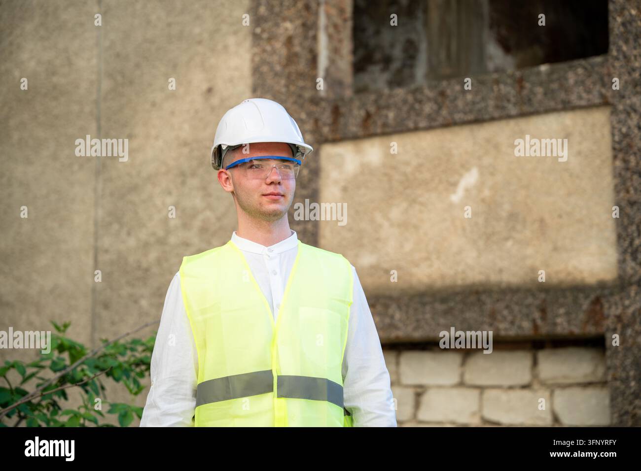 Professionista dell'edilizia con casco protettivo e giubbotto ad alta visibilità accanto a un edificio esterno incompiuto. Assunzione di operai qualificati architettura un Foto Stock