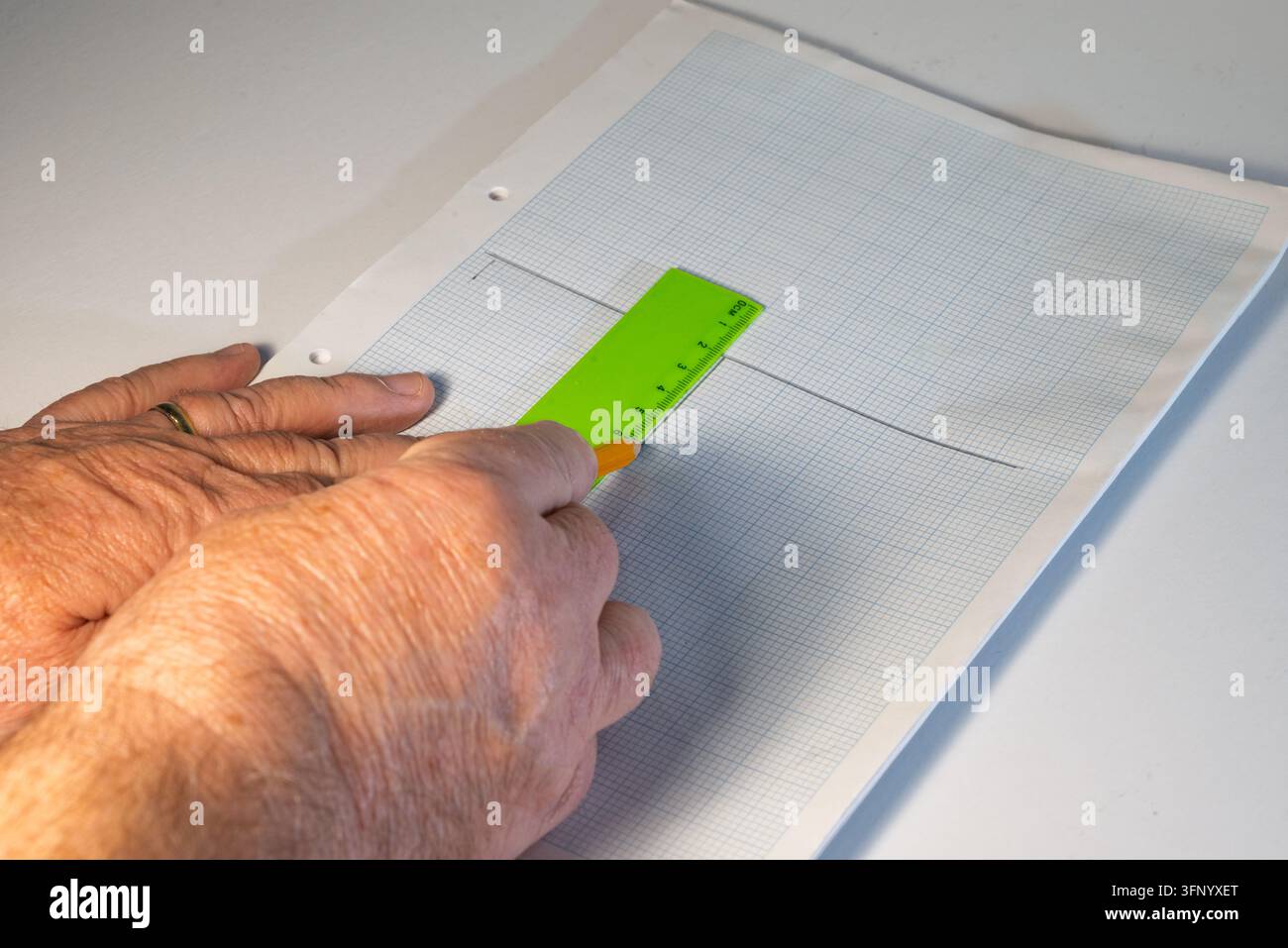Mani adulte tracciano una linea su carta millimetrata usando un righello verde e una matita gialla. Un anello d'oro è visibile su un dito. Foto Stock