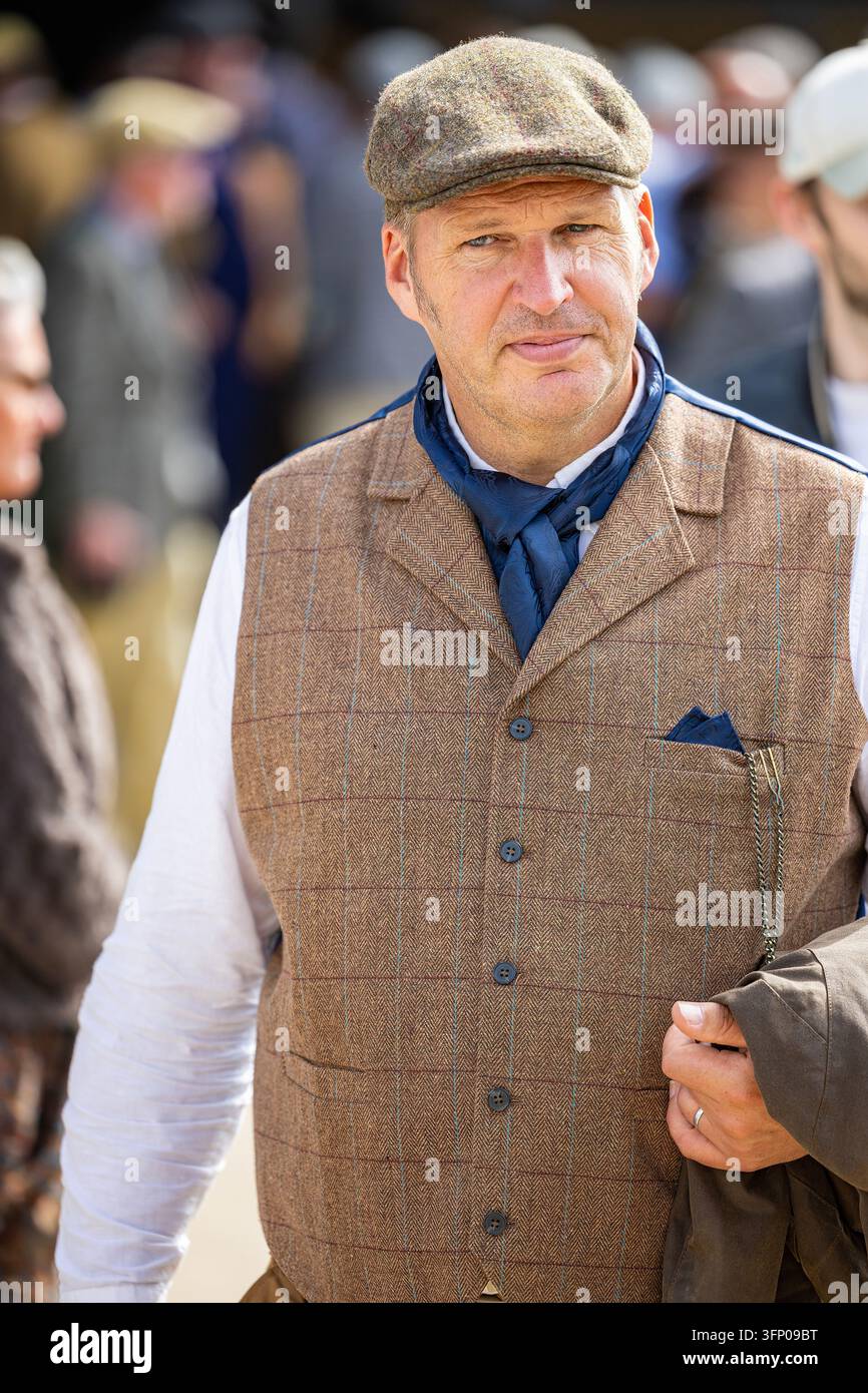 Partecipante al Goodwood Revival in gilet di tweed, berretto piatto e foulard che porta una giacca tra la folla del paddock. Foto Stock