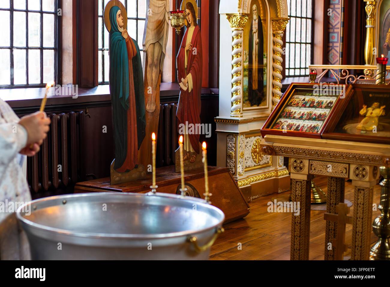 Una persona tiene una sottile candela accesa vicino a un bacino d'acqua santa d'argento all'interno di un tempio ortodosso. Rito cristiano di battesimo sacramento cerimonia Foto Stock