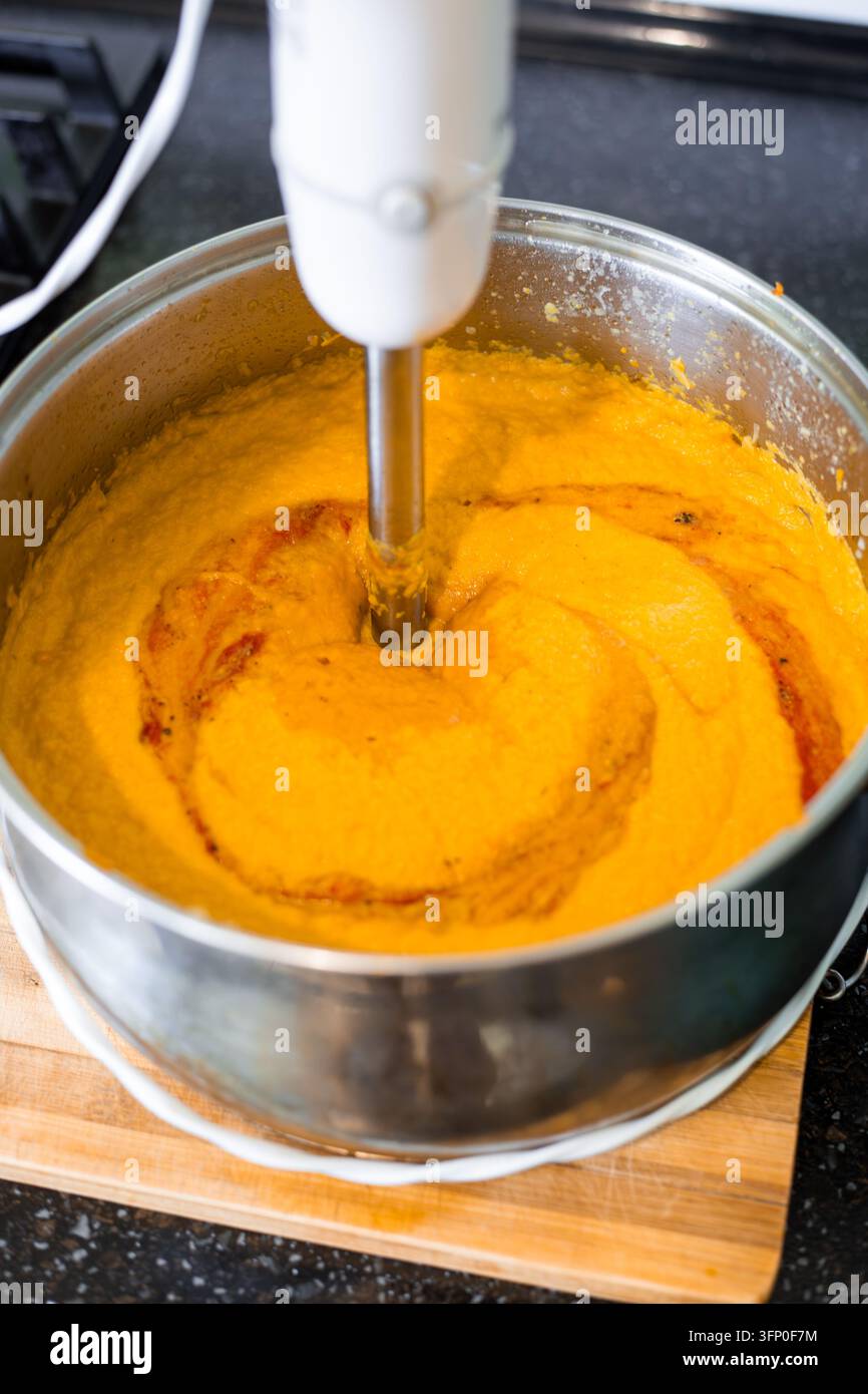 Un frullatore elettrico a mano bianco riduce in purea una zuppa di verdure arancione fresca dentro una pentola di metallo. Processo di cottura del zucca fatto in casa. Tema alimentare, design culinario. Foto Stock