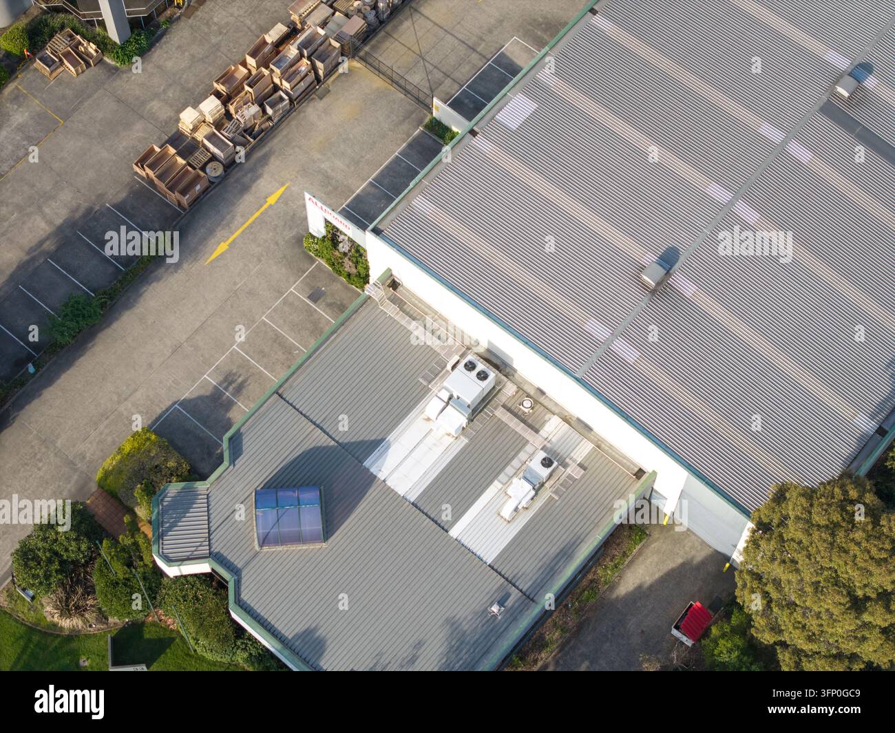 Vista aerea di un edificio industriale con tetto in lamiera ondulata, unità HVAC e lucernari, adiacente a un'area di parcheggio con segnaletica gialla e legno Foto Stock