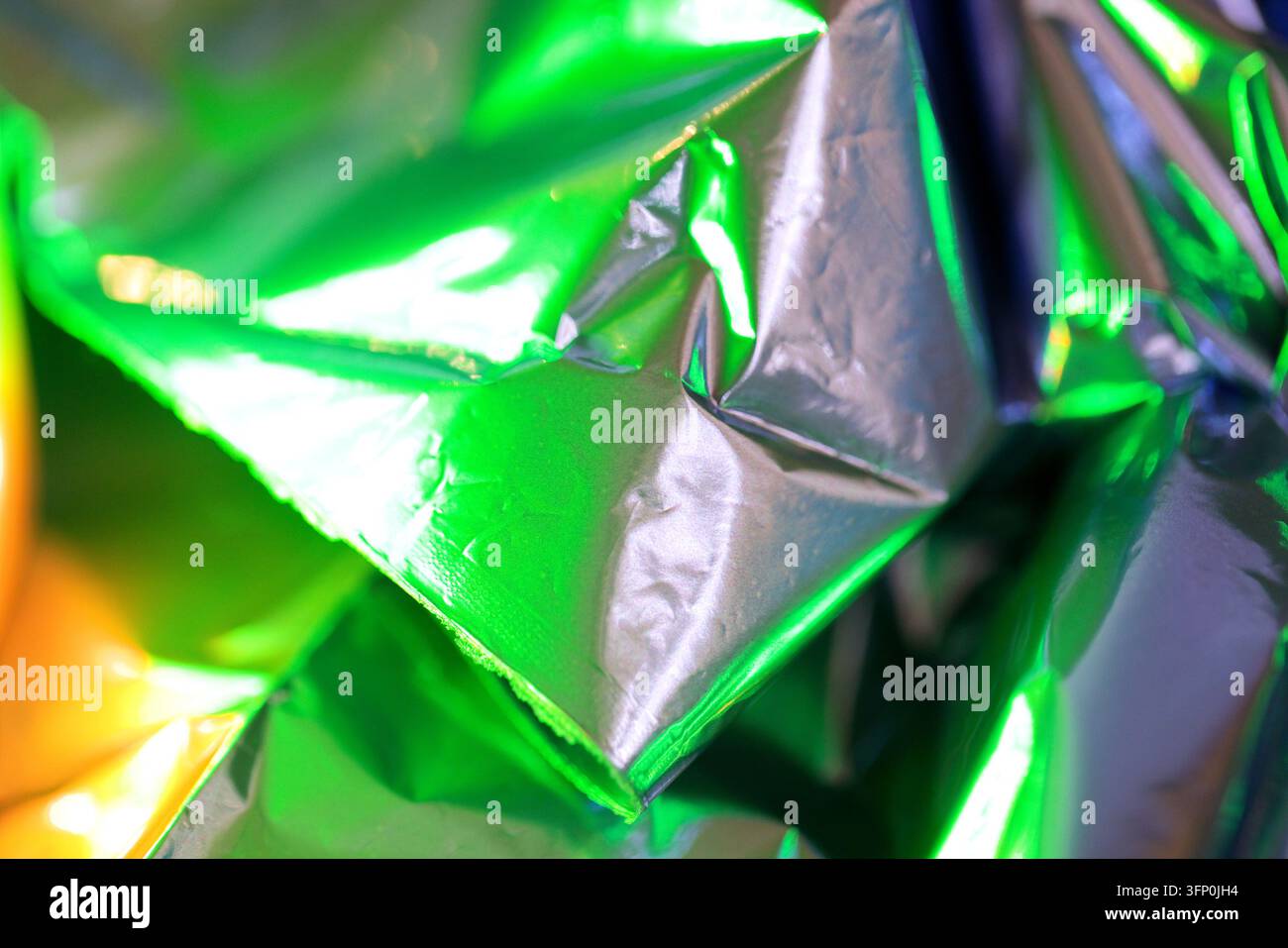Foglio riflettente raggrinzito forma pieghe angolari nette attraverso il quadro, catturando una luce vivace verde e gialla in una texture metallica astratta e lucida Foto Stock