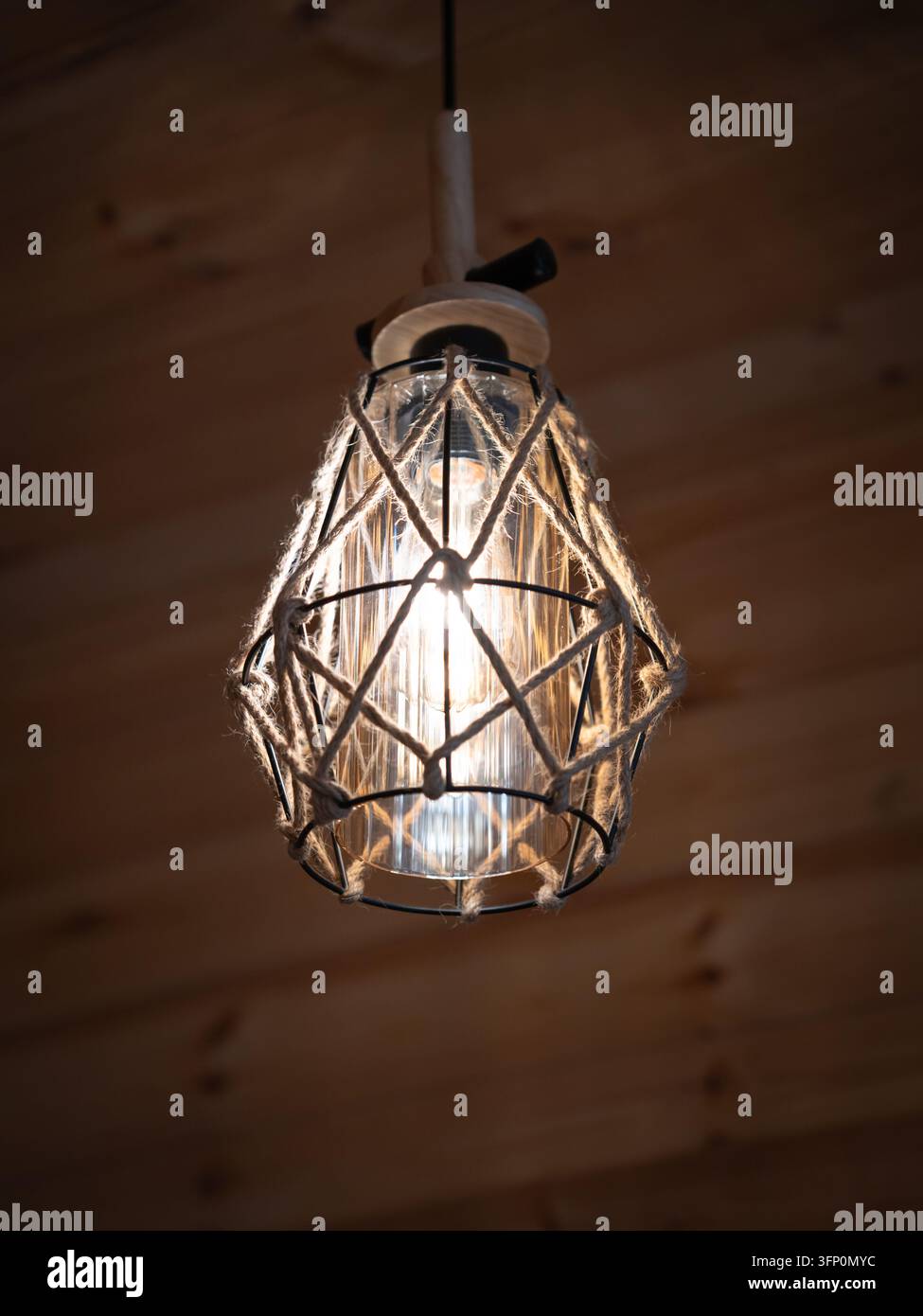 Lampadario a sospensione con bulbo Edison e gabbia decorativa in corda che diffonde una luce calda, esaltando il design interno accogliente e rustico di un soffitto con pannelli di legno. Ver Foto Stock