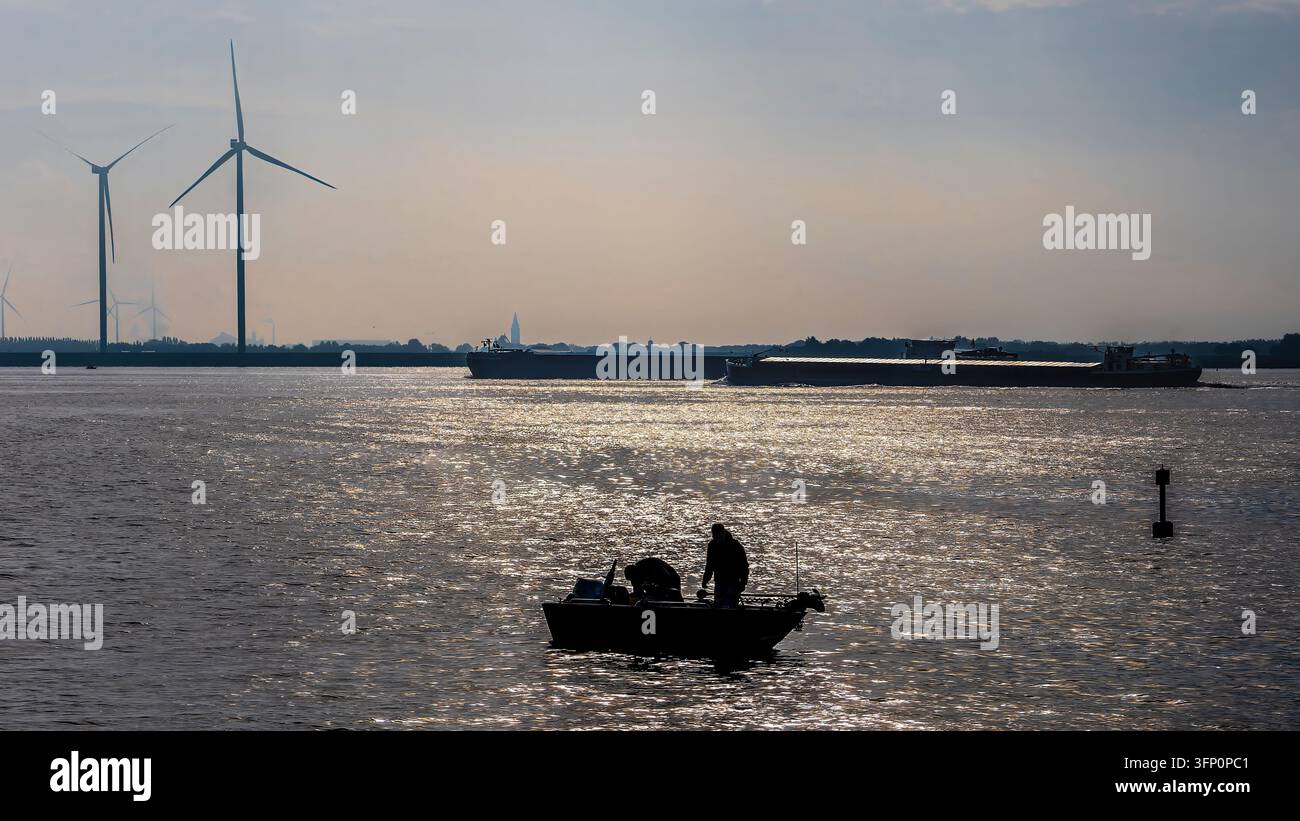 Pescatore su una barca che si gode il sole del tardo pomeriggio sull'acqua. Mentre grandi chiatte da carico navigano il fiume. E turbine eoliche generano energia pulita sullo sfondo vicino a Ooltgensplaat. Paesi Bassi Foto Stock